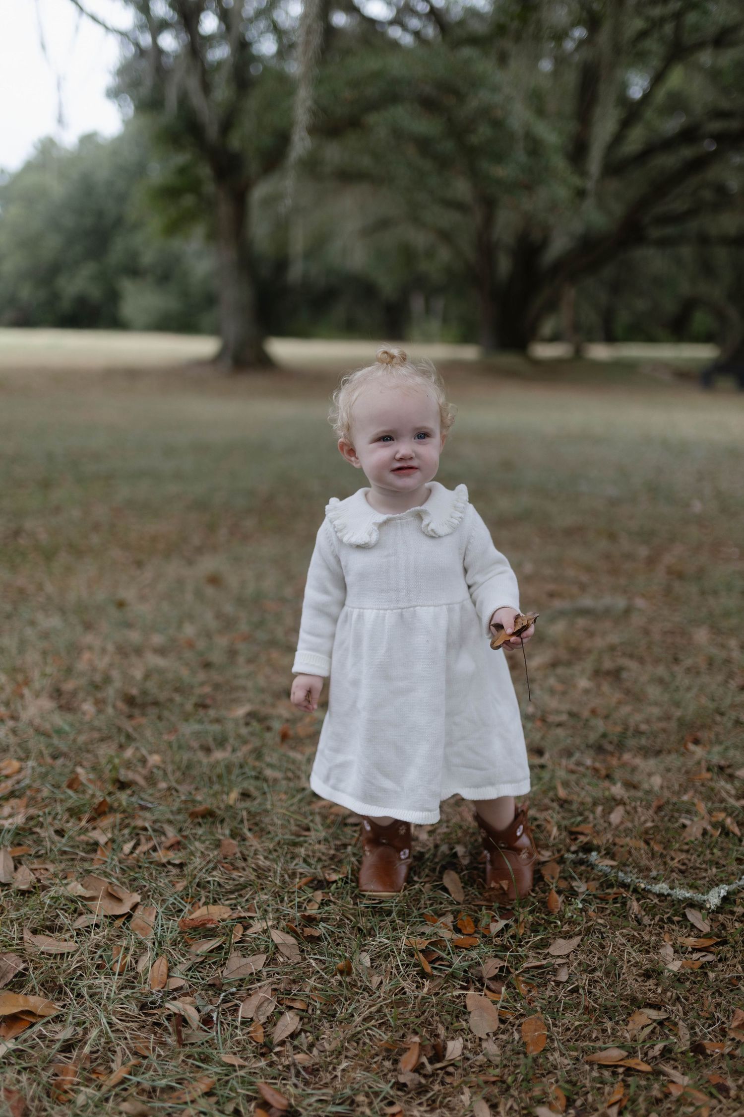 Small child in white dress stands among fallen autumn leaves in a park setting.