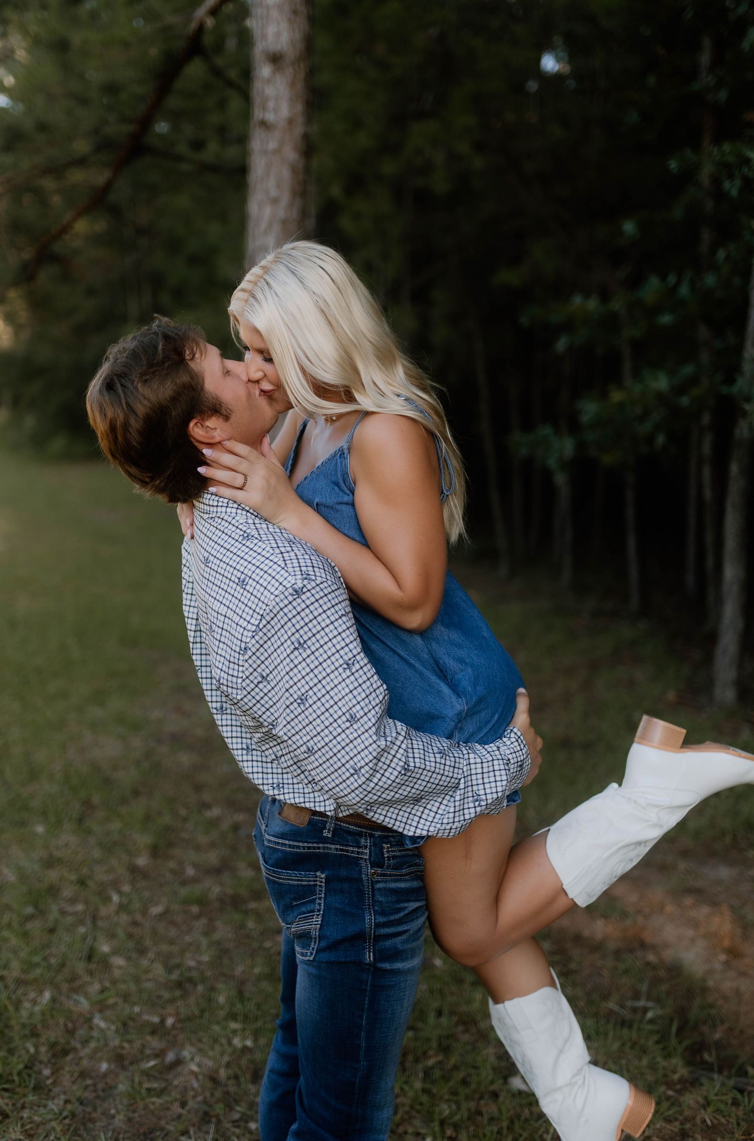 A couple in blue and plaid share romantic kisses outdoors in a wooded setting during a photo session.