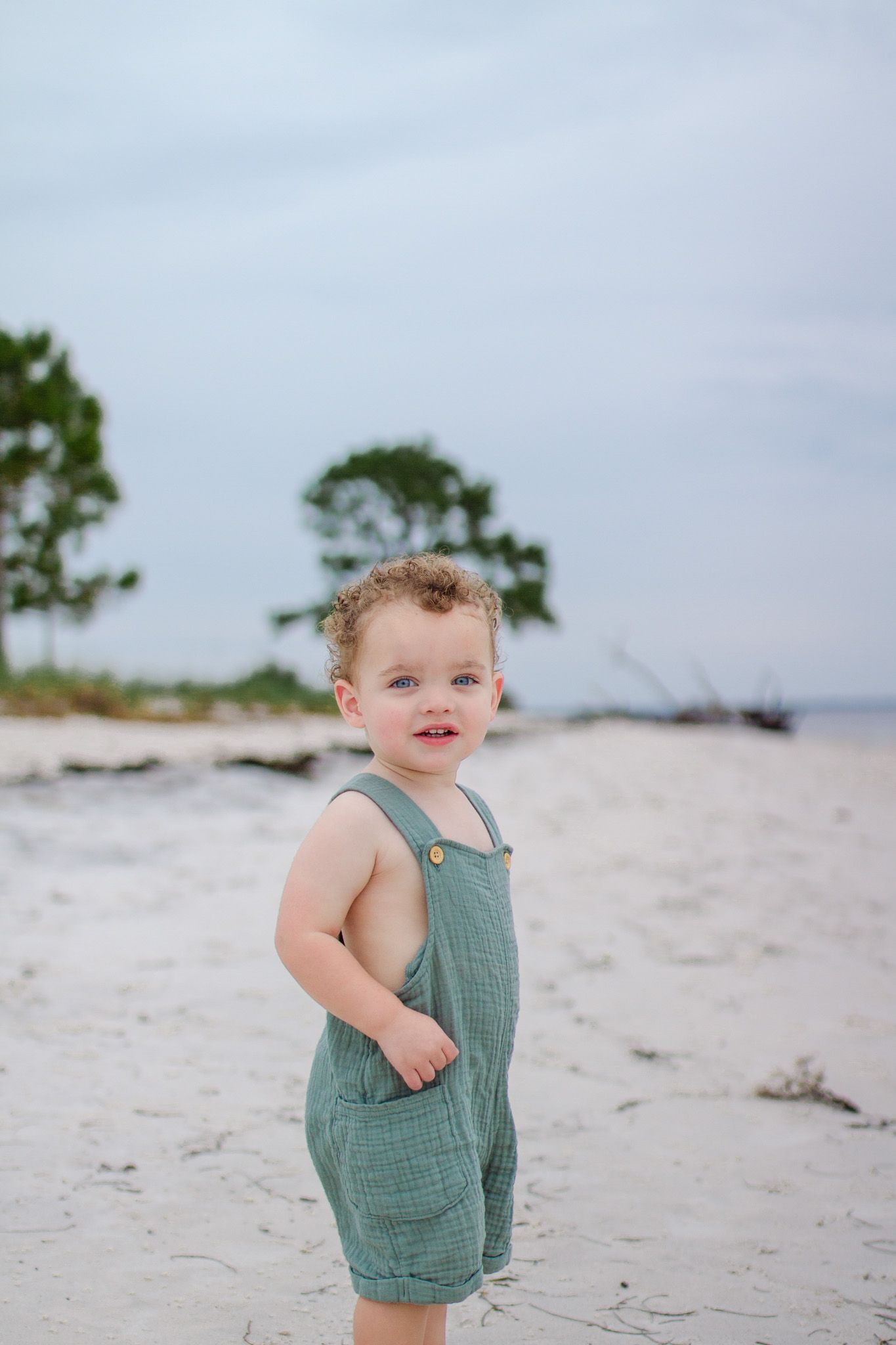 A toddler wearing a teal linen romper stands on a sandy beach with pine trees in the background.