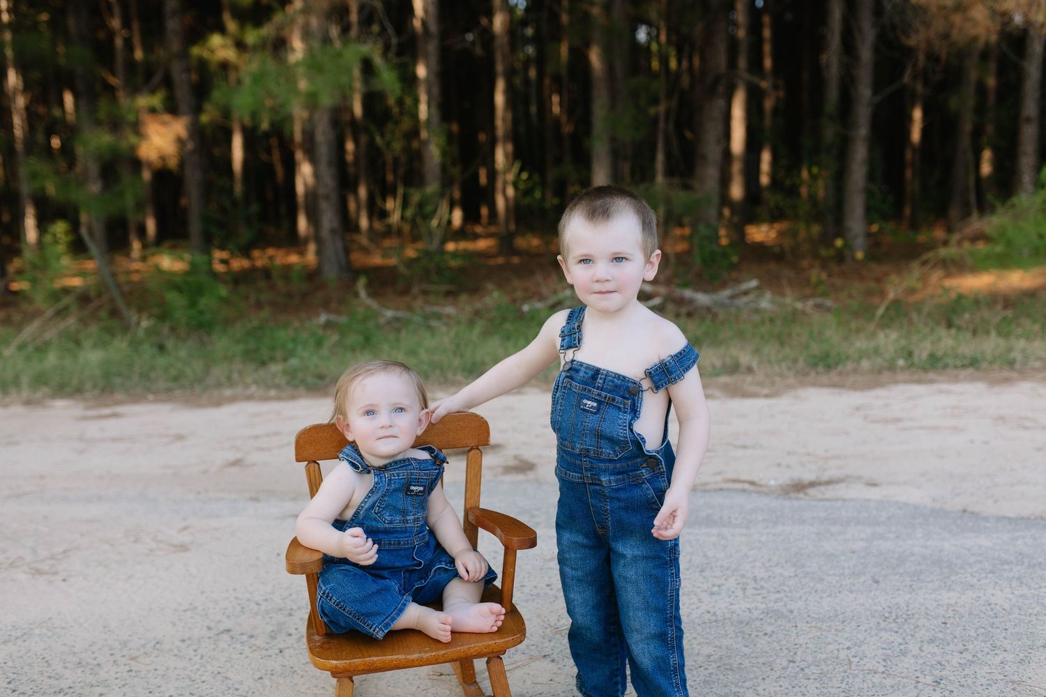 A child sits in a wooden chair on a gravel path while someone in denim stands nearby.