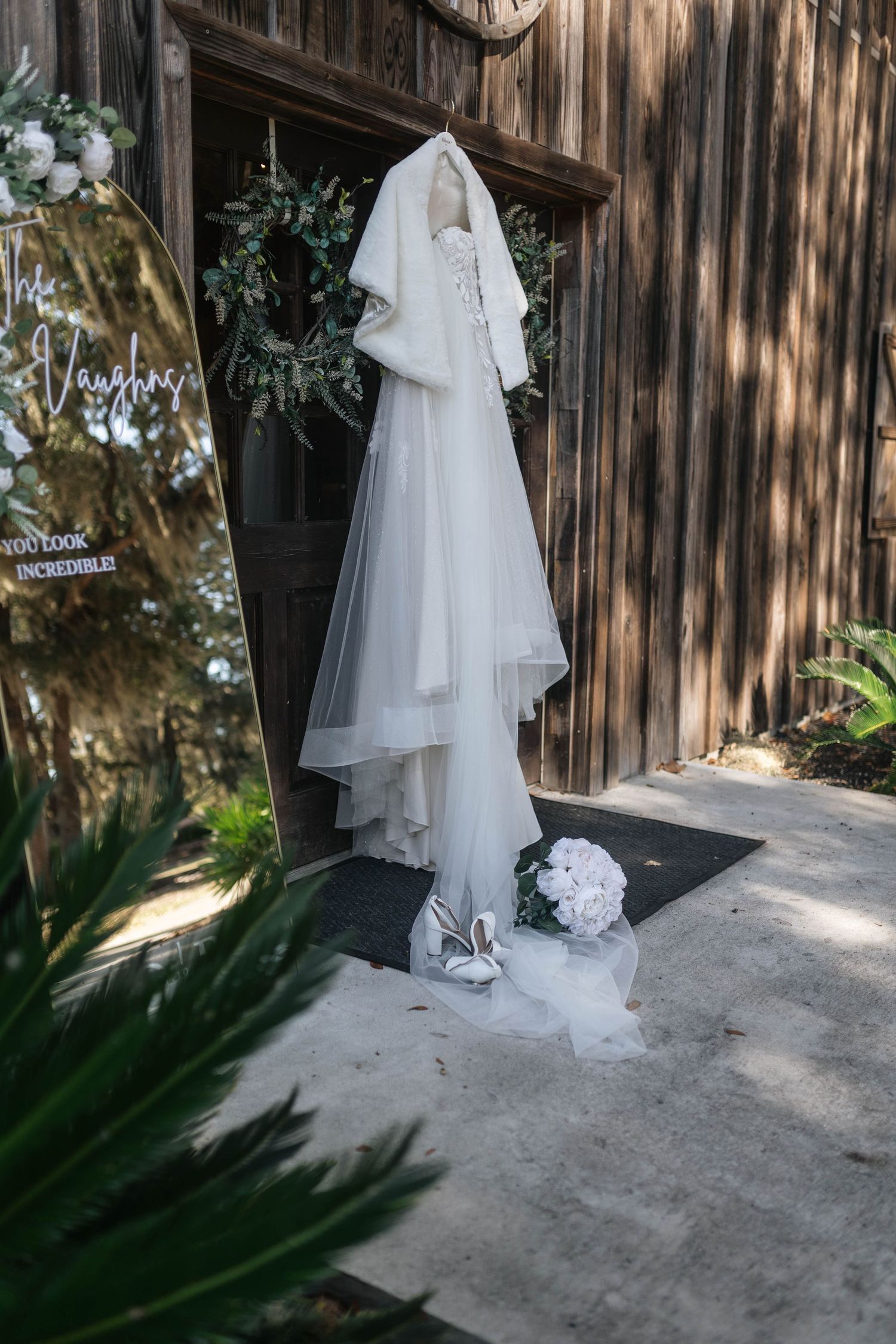 A white wedding dress and veil hanging on a rustic wooden wall with palm fronds visible in the foreground.