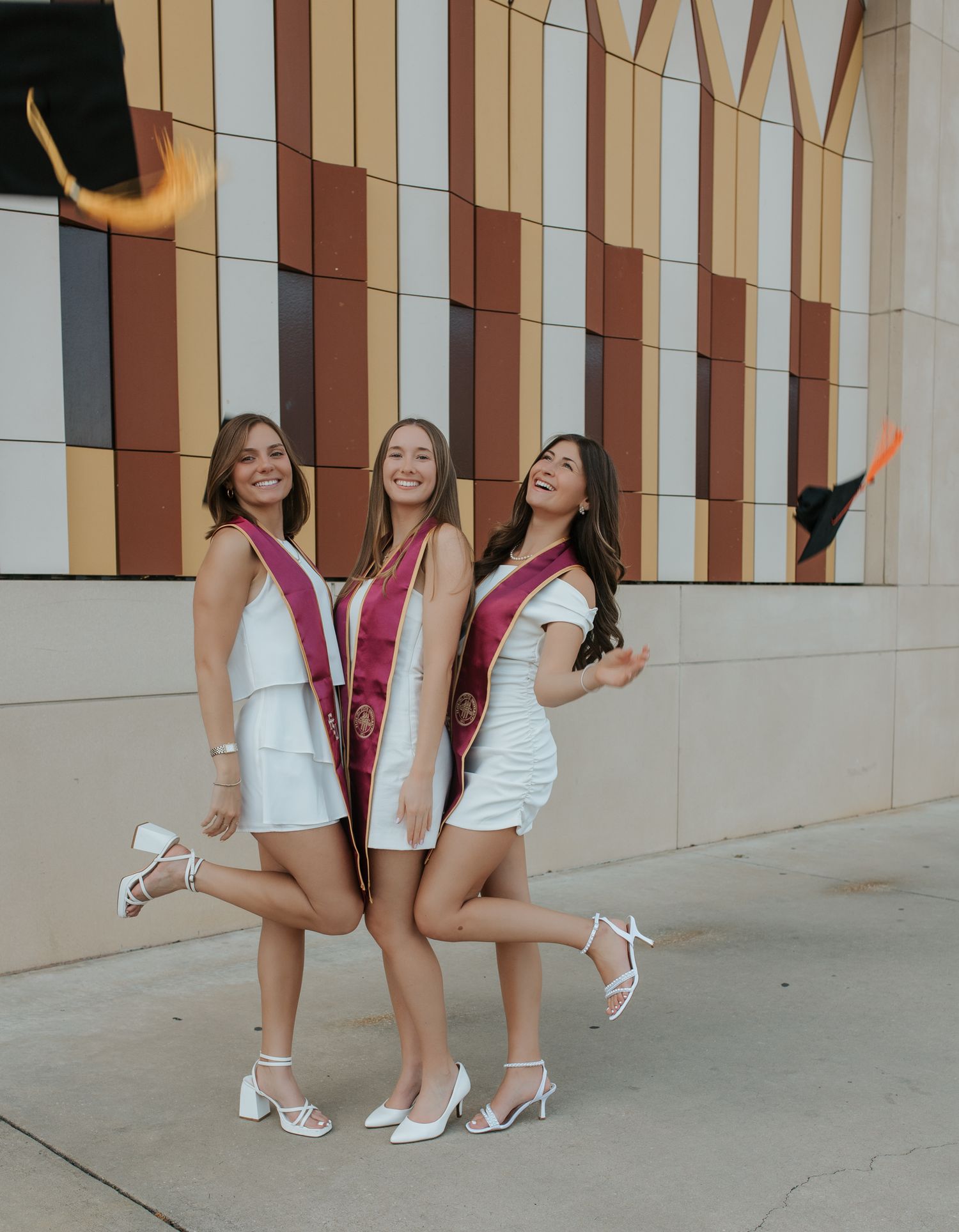 Group of friends in white dresses and burgundy jackets pose playfully in front of a striped wall.