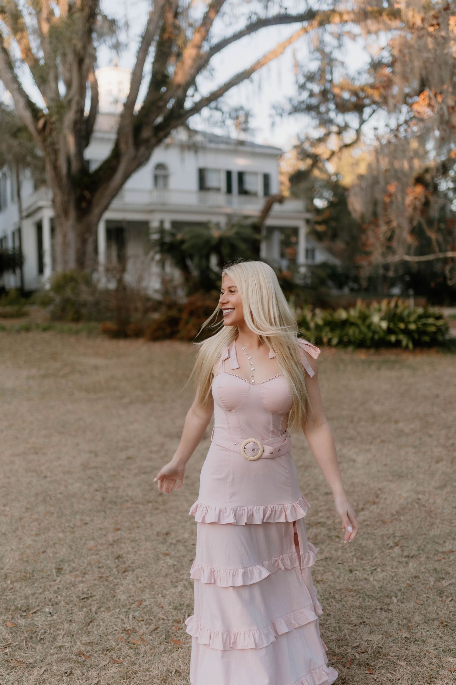 A person wearing a pink ruffled dress twirls in an autumn yard with bare trees and white houses in the background.