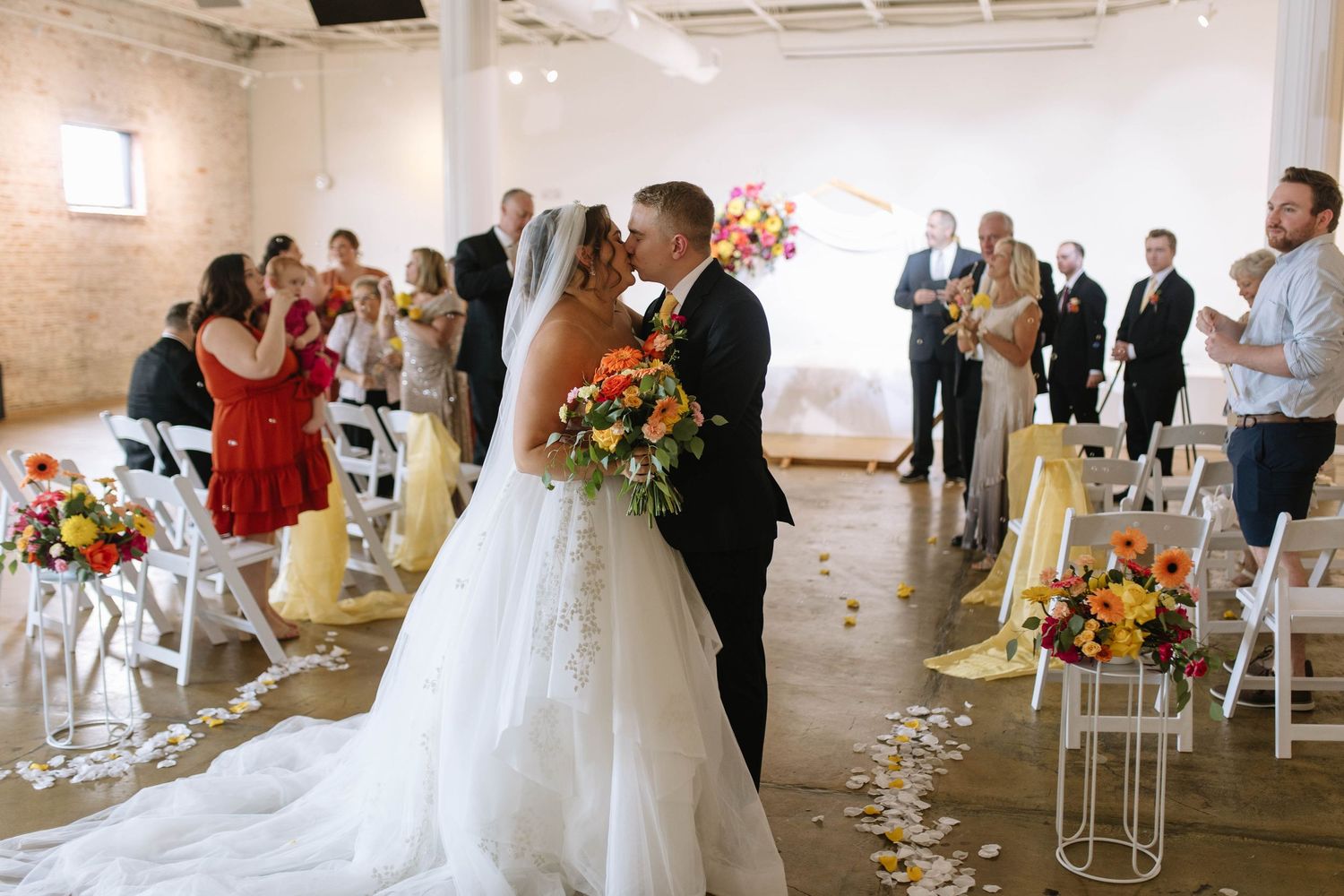 A wedding ceremony in a bright indoor venue with white chairs and colorful floral arrangements along the aisle.