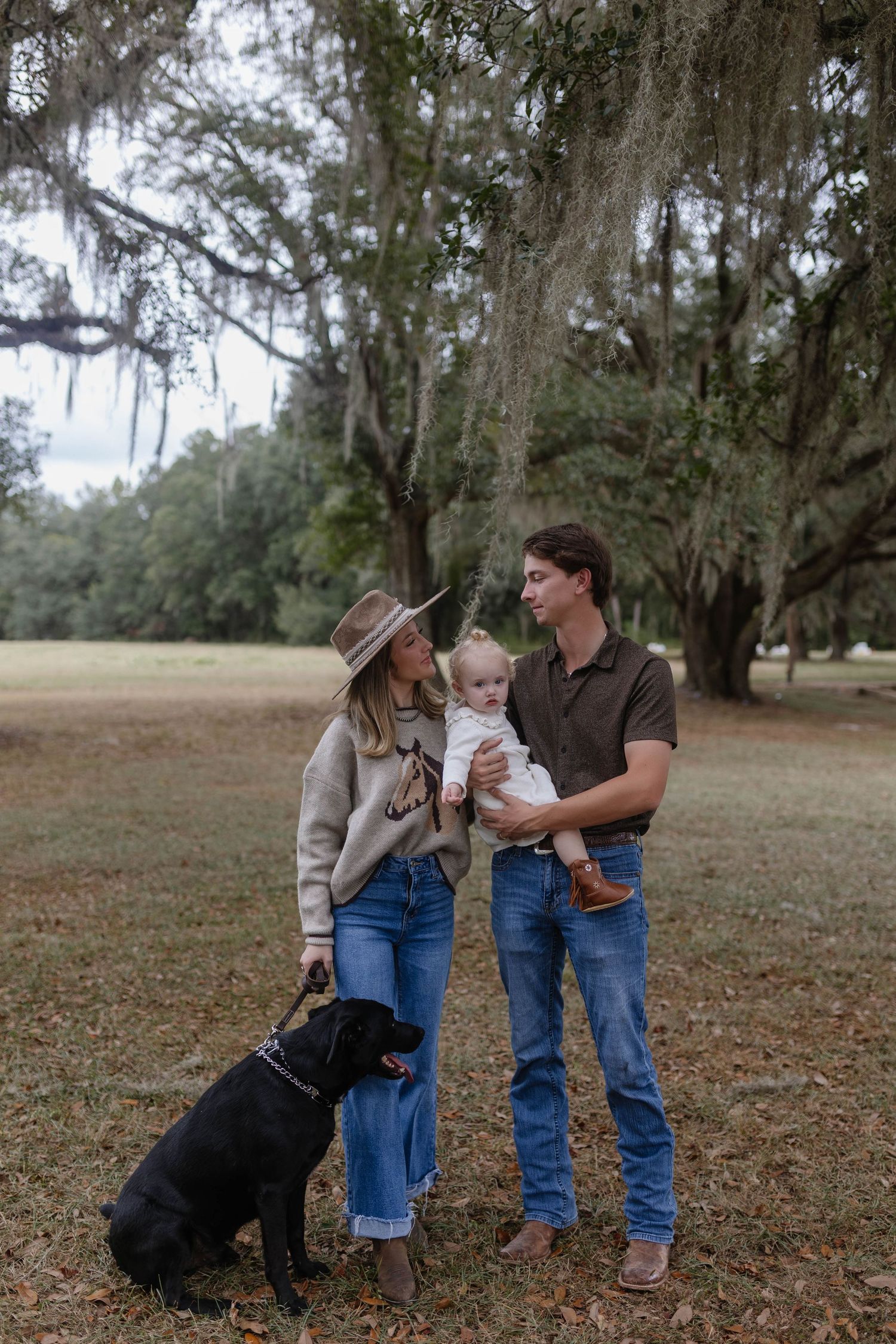 A family and their black dog share a tender moment together on fallen autumn leaves in a park setting.