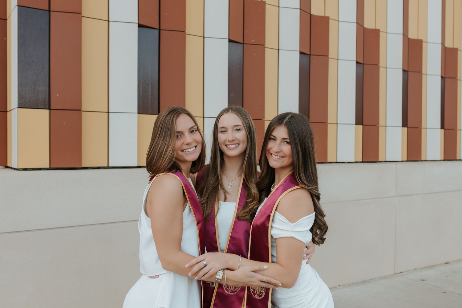 Three graduates in white dresses and red graduation stoles pose together in front of a modern building wall.