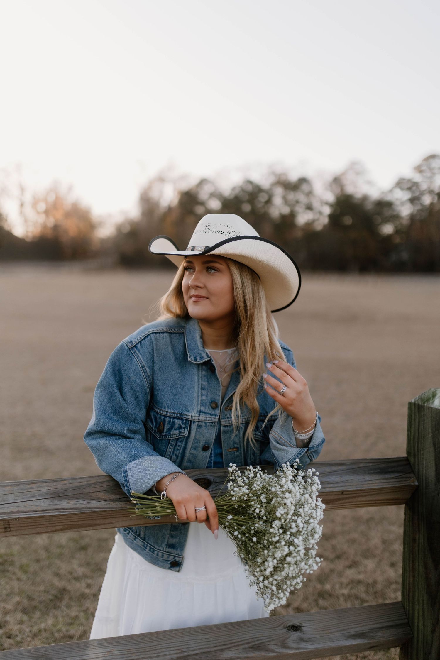 Woman in denim jacket and white dress sits on wooden fence holding baby's breath flowers at sunset.