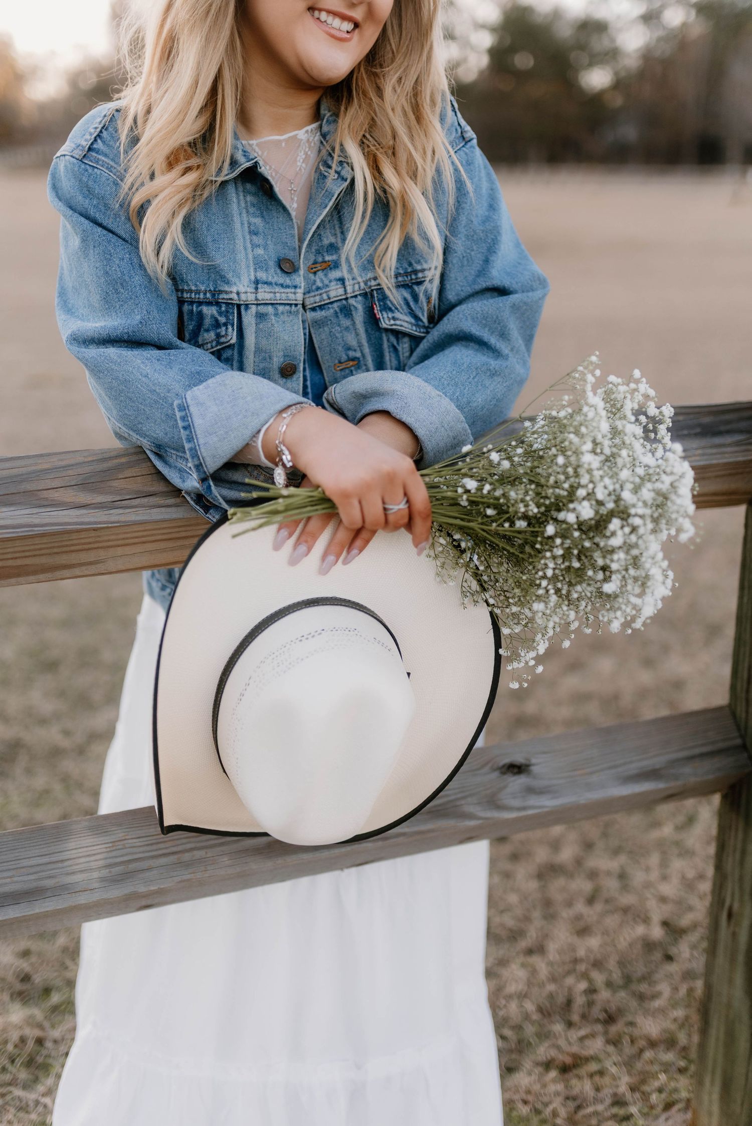 A white sun hat with baby's breath flowers rests on a wooden bench while someone in denim jacket holds it.