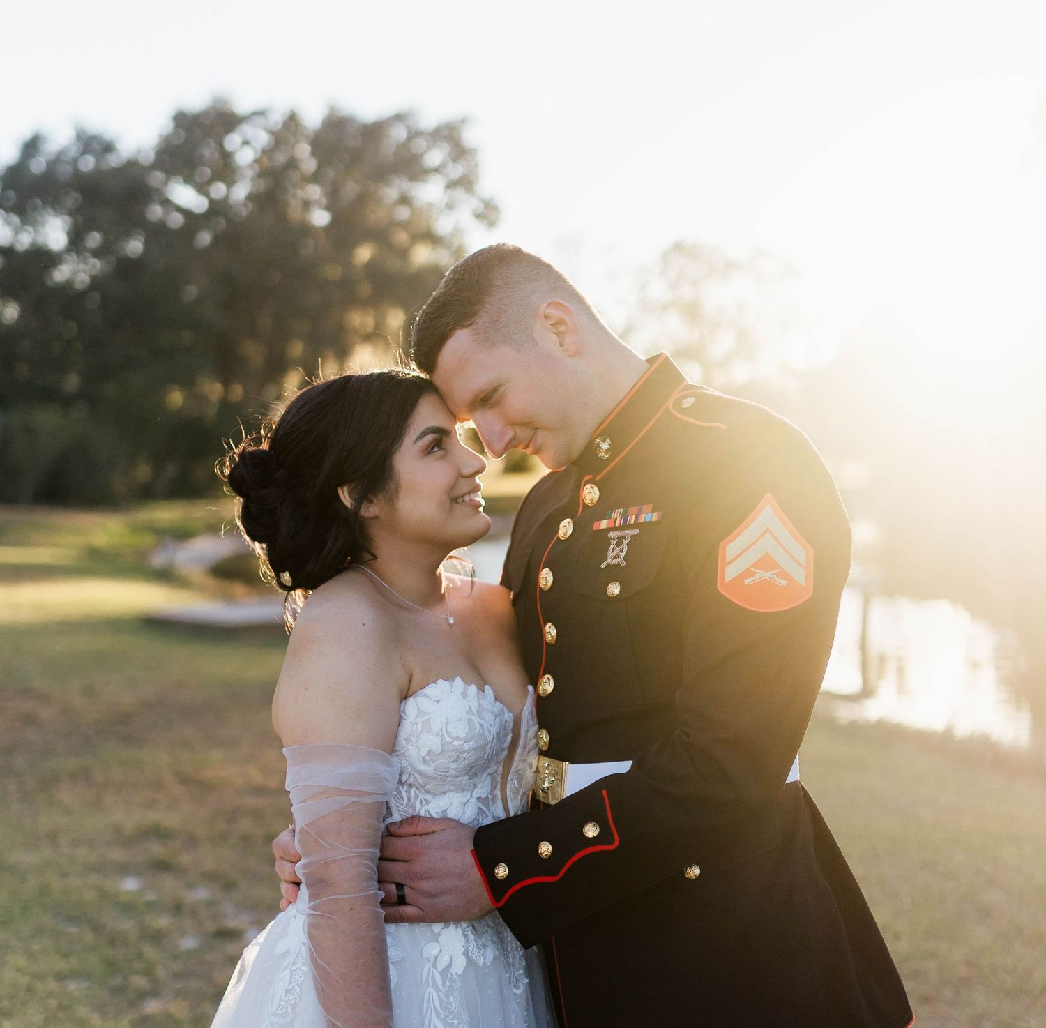 A couple in formal wedding attire share an intimate moment at sunset in a scenic outdoor setting.