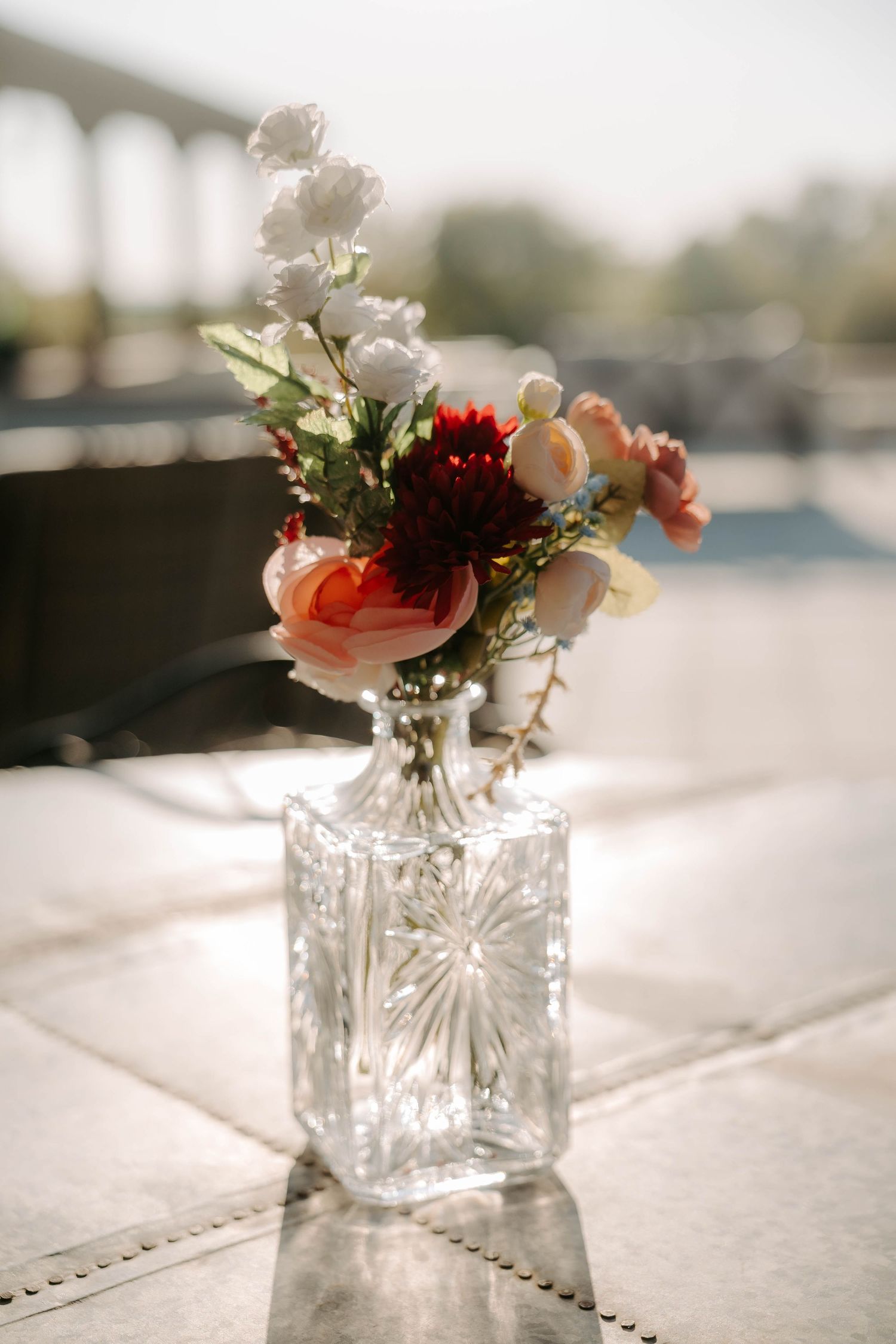 Crystal vase with red, pink, and white flowers on a sunlit table outdoors.