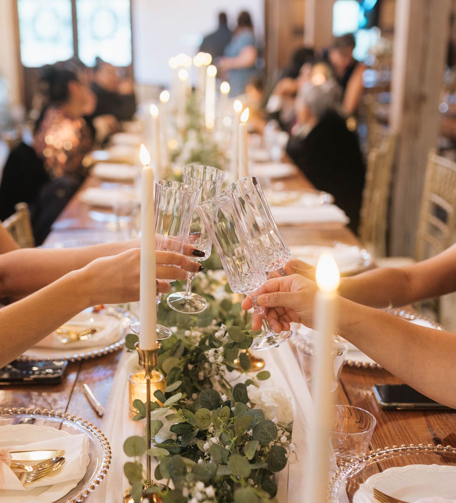 An elegant wedding table setting with white candles, greenery garlands, and crystal glasses in a softly lit reception venue.