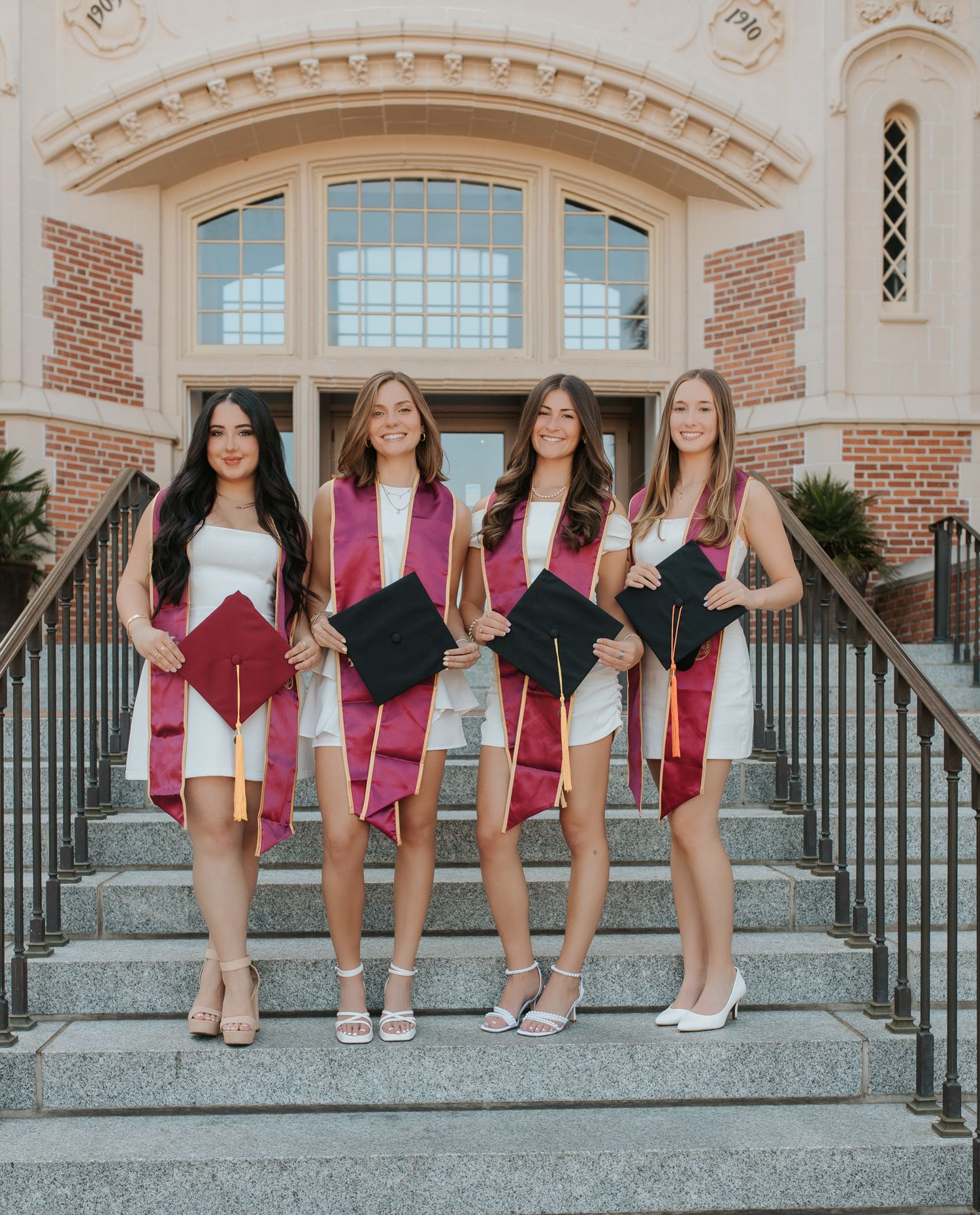 A group of friends in pink dresses sit together on stone steps in front of a brick building entrance.