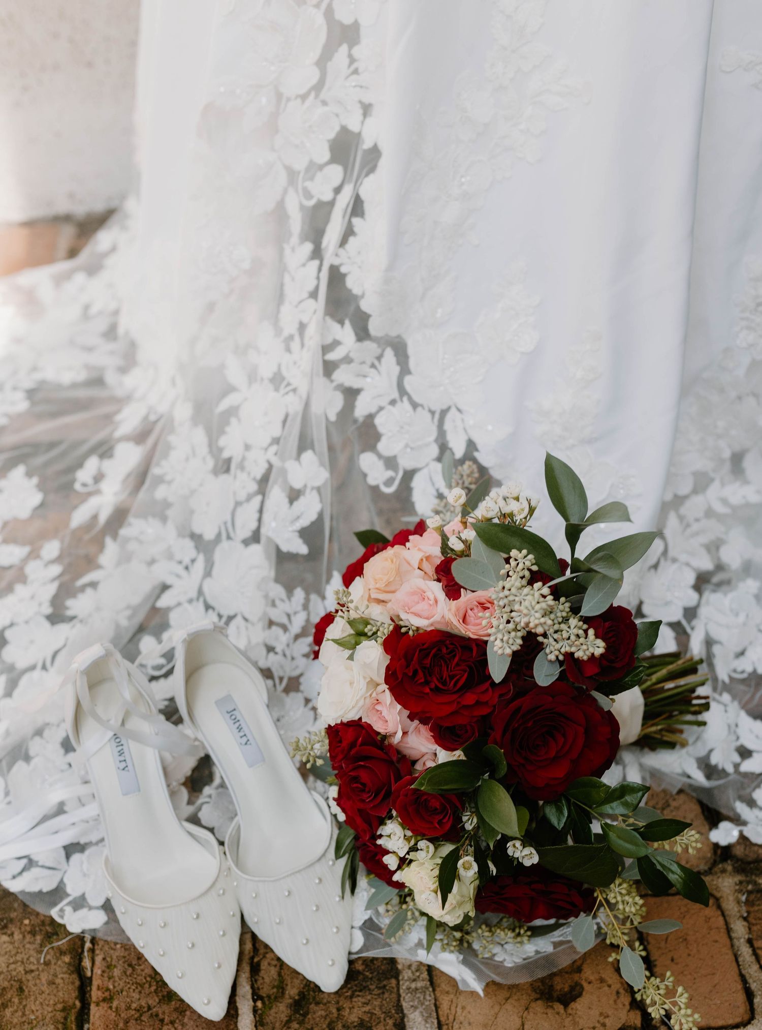 Bridal flat shoes, red and pink rose bouquet, and lace wedding dress train arranged on rustic brick floor.