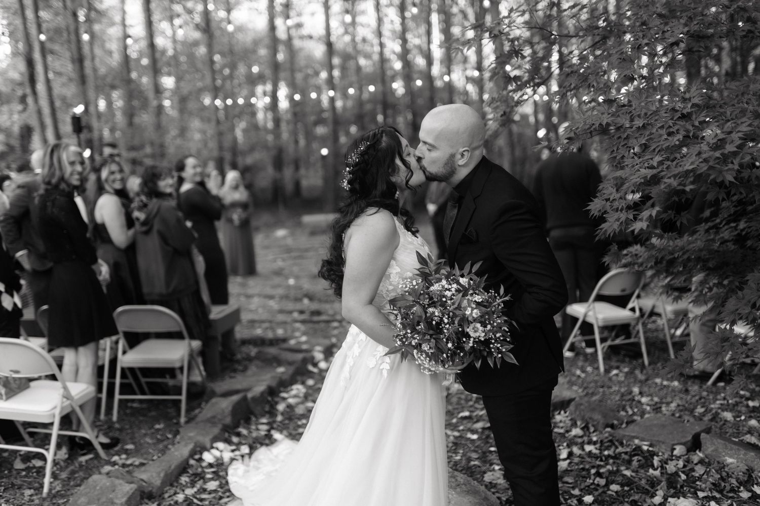 A black and white wedding ceremony takes place in a serene forest setting with guests seated on folding chairs.