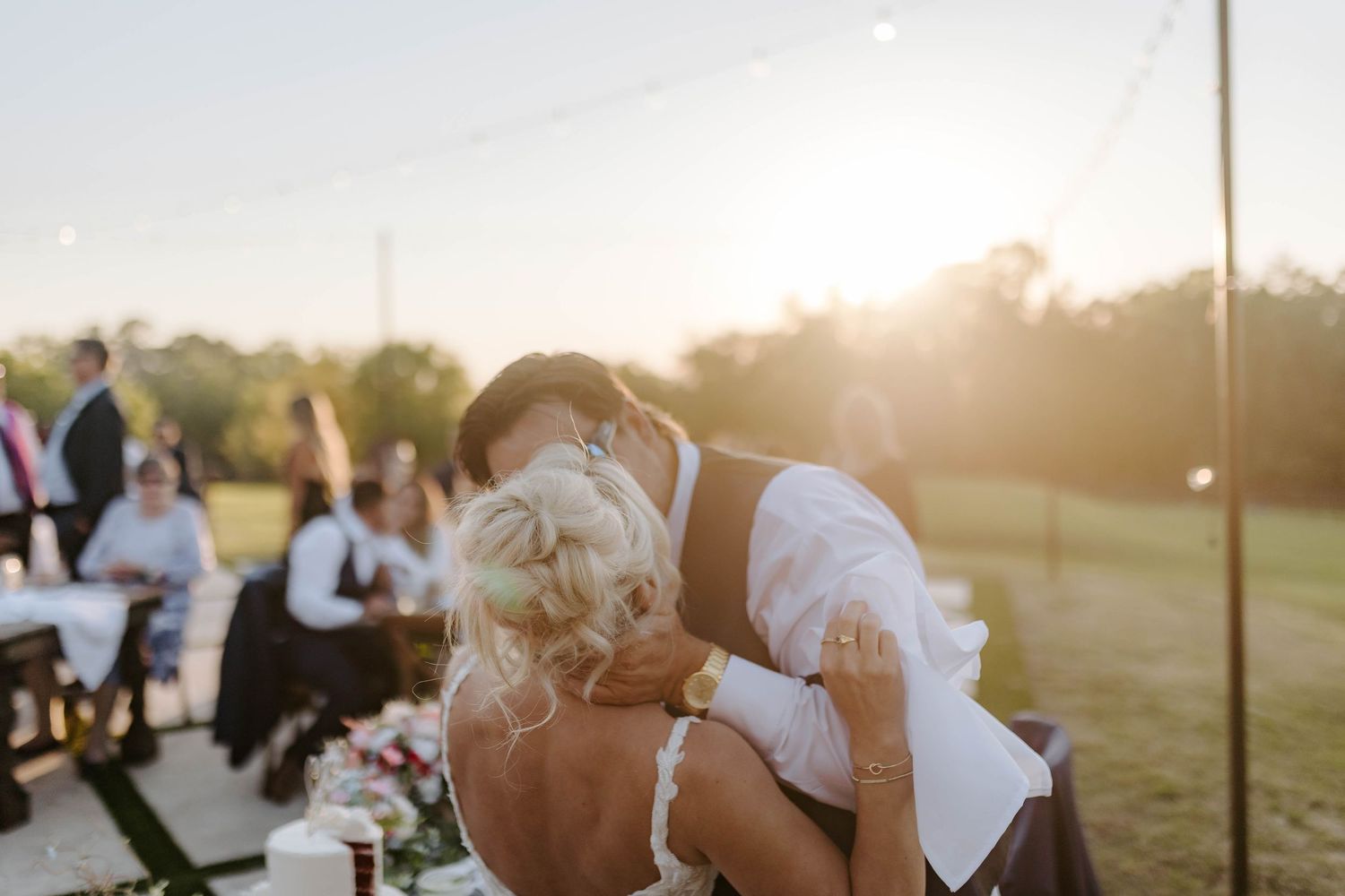 Bride and groom share a romantic kiss at golden hour outdoor wedding reception with guests seated in background.