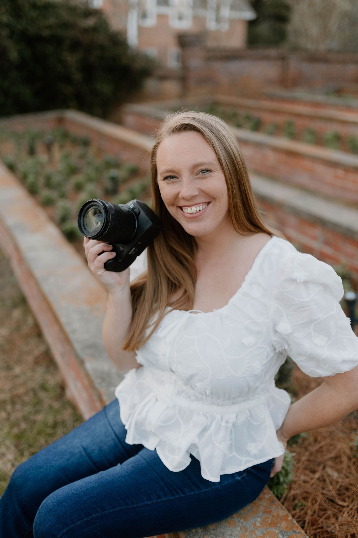 A photographer in a white ruffled top and jeans sits outdoors holding a professional camera with a telephoto lens.