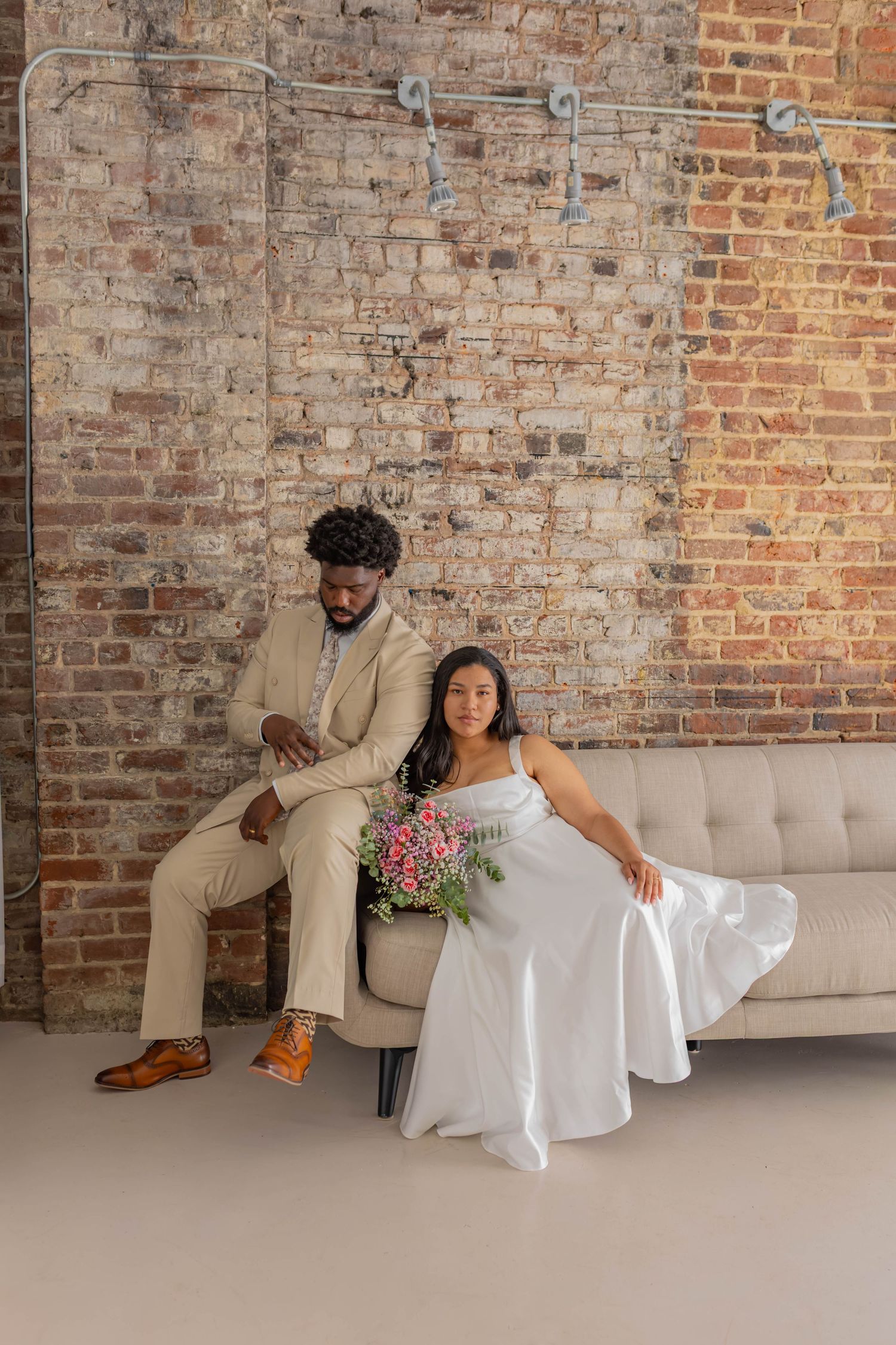 Bride in white gown holds pink bouquet beside groom in tan suit, seated on gray sofa against rustic brick wall.