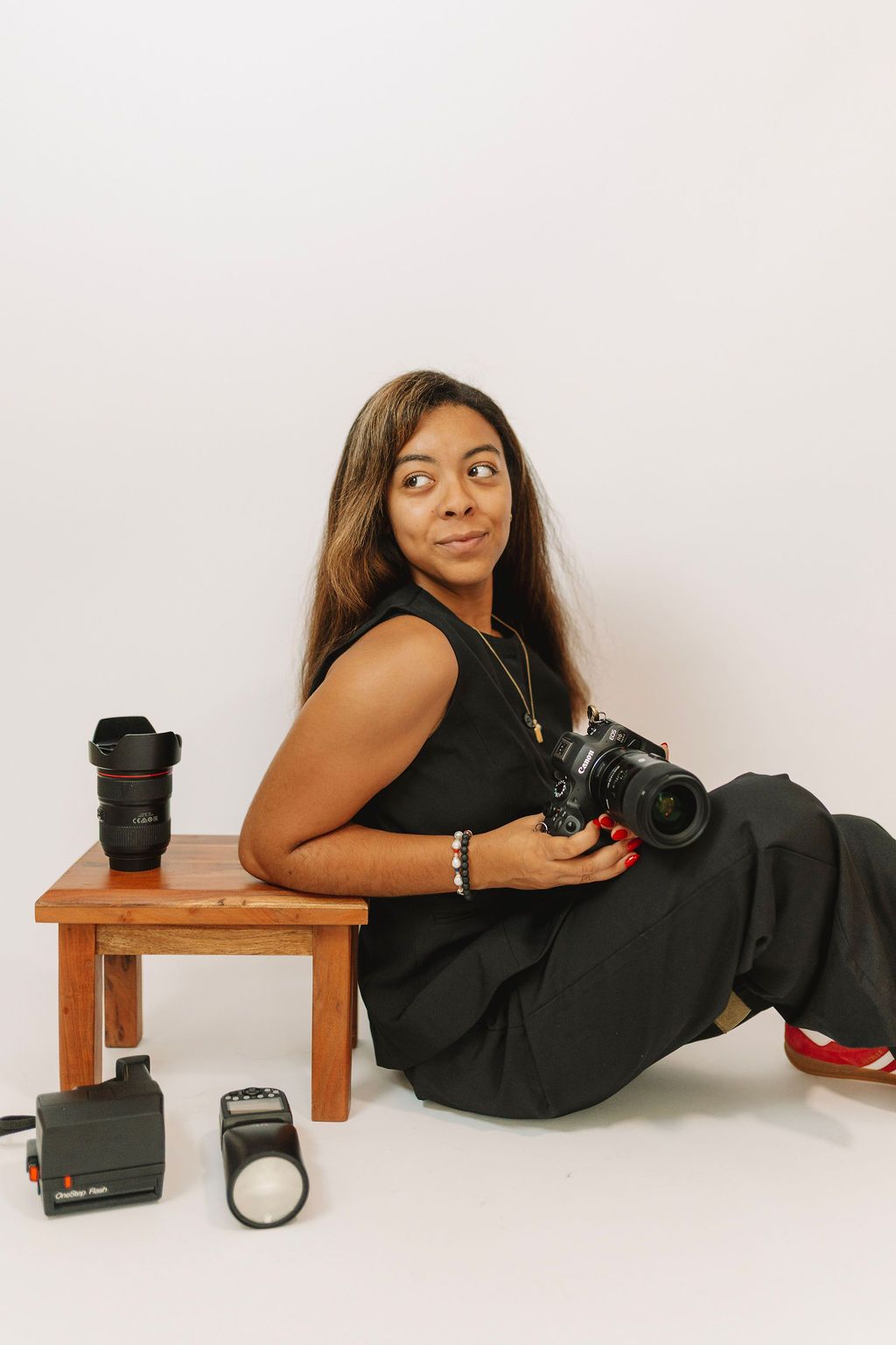 Woman photographer sitting on floor holding DSLR camera with lens and flash equipment beside a wooden stool.
