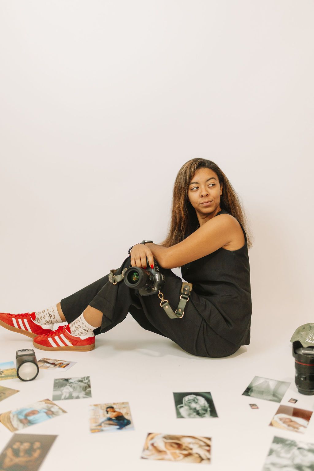 Young woman photographer sitting on studio floor with camera, surrounded by printed photos, wearing red Adidas sneakers and black outfit.
