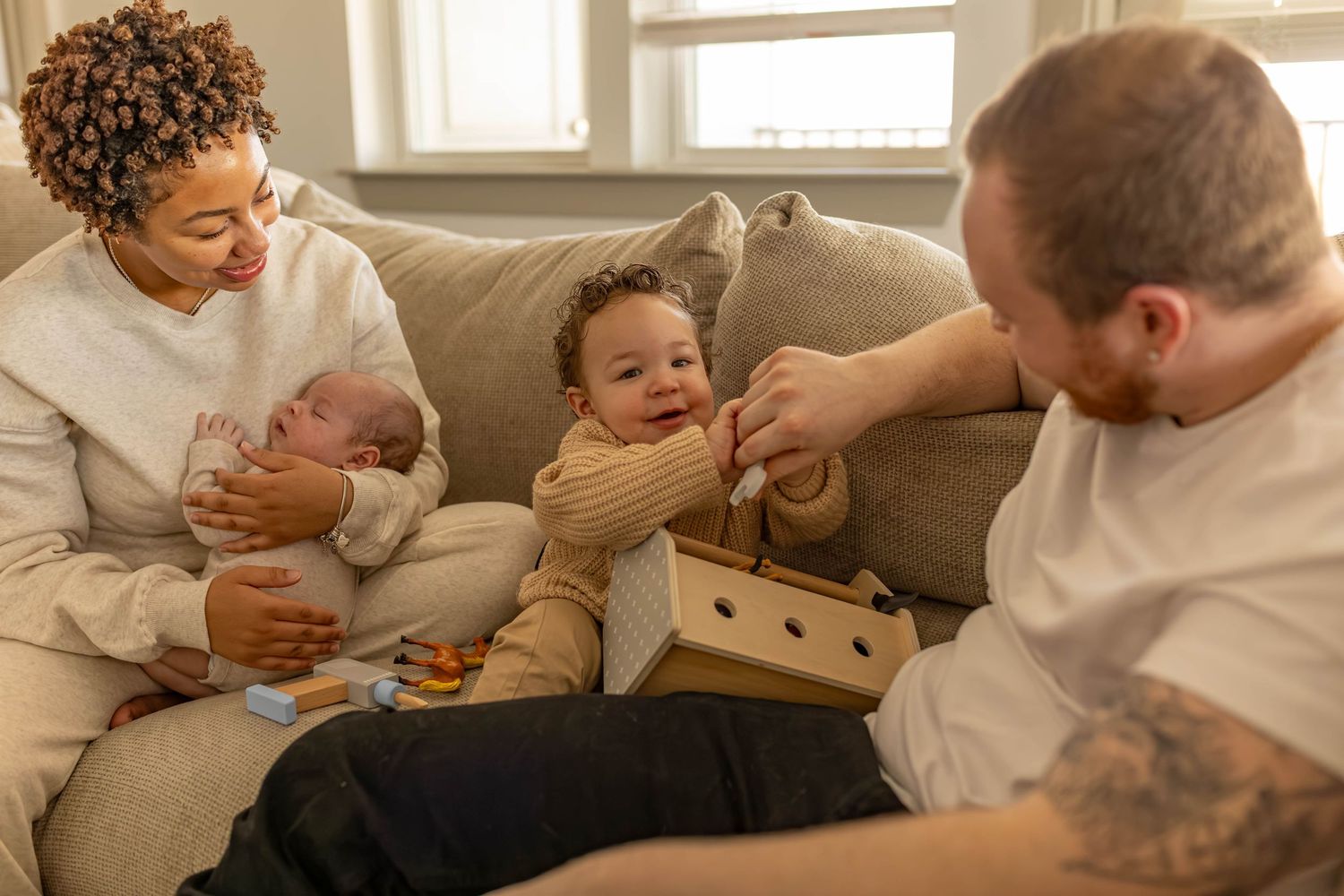 Happy family with two young children sitting on a couch, parents playing with toddler while holding newborn baby.