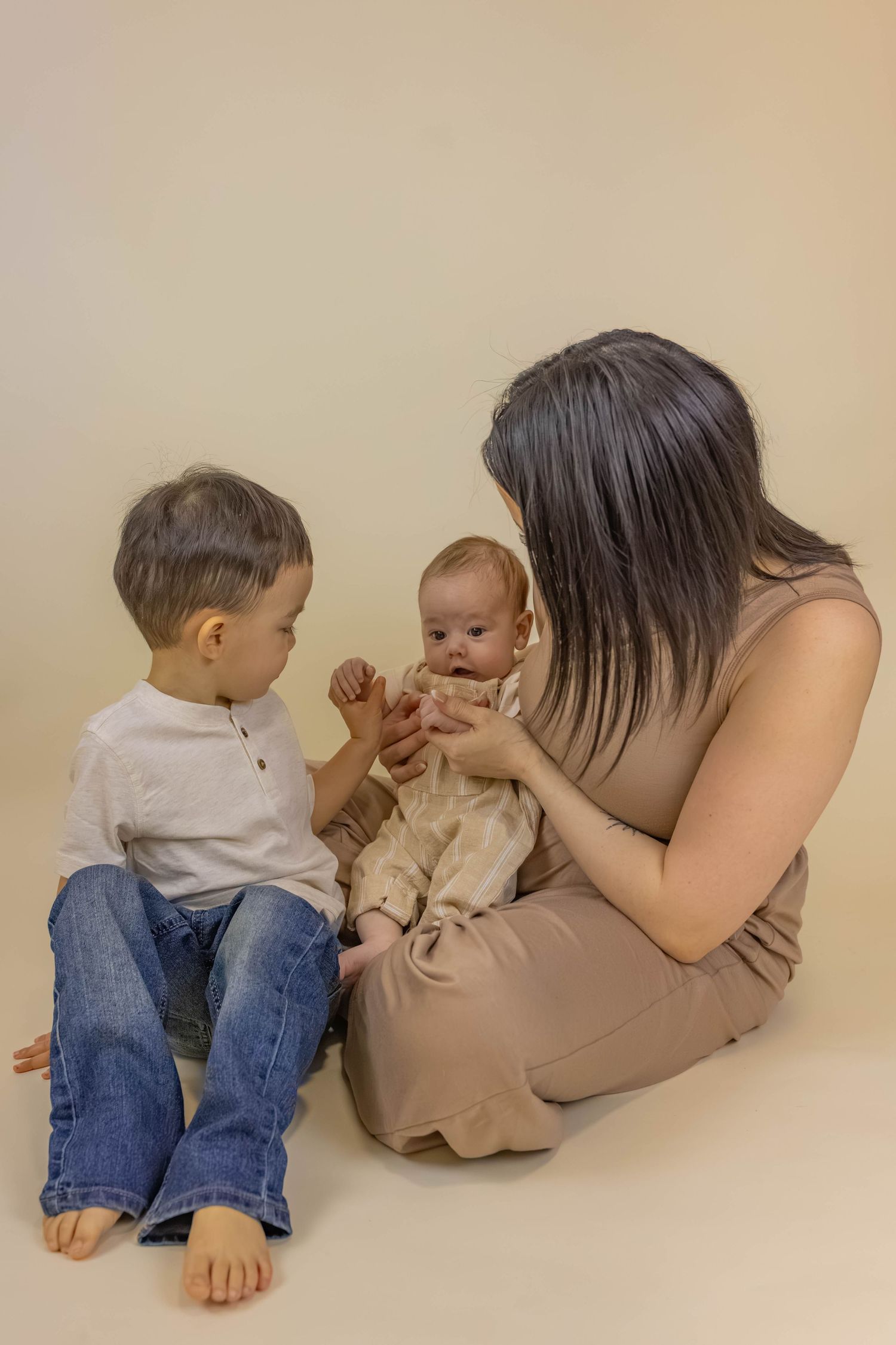 A young mother sits on the floor holding a newborn baby while her toddler son in jeans looks on curiously.