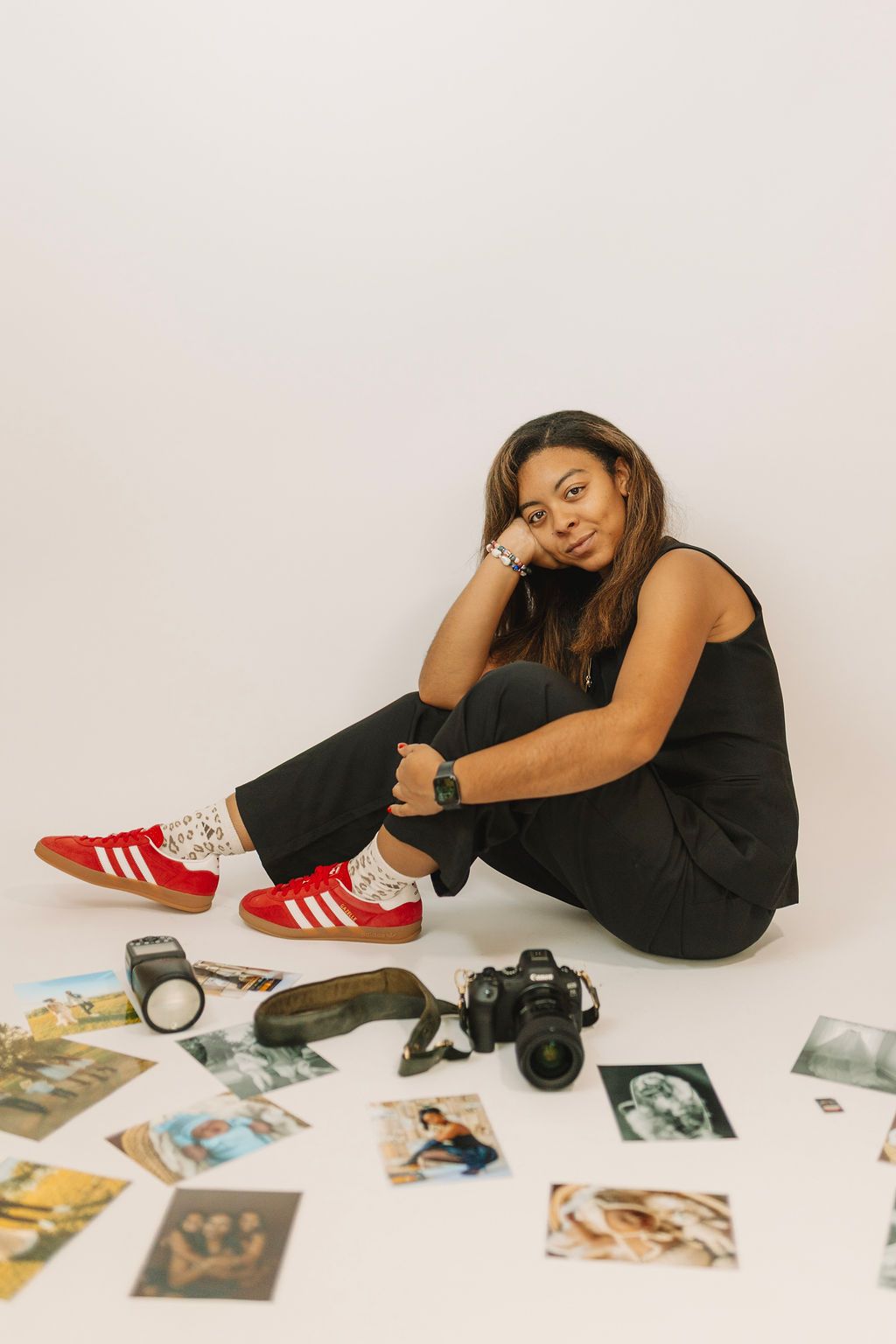 Woman in black outfit and red Adidas sneakers sits on white floor surrounded by photos, camera, and photography equipment.