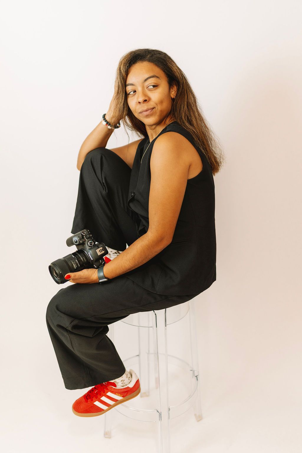 Woman in black outfit holding a DSLR camera, seated on a clear stool, wearing red sneakers against white background.
