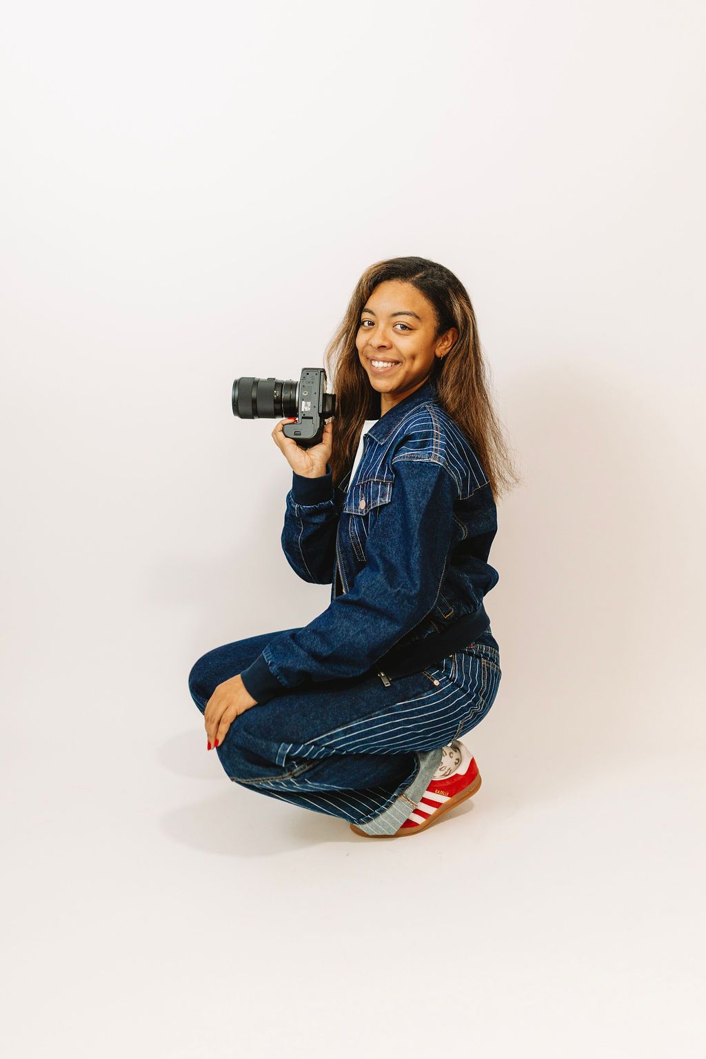 Young woman in denim outfit crouching while holding a camera, smiling against a white background.