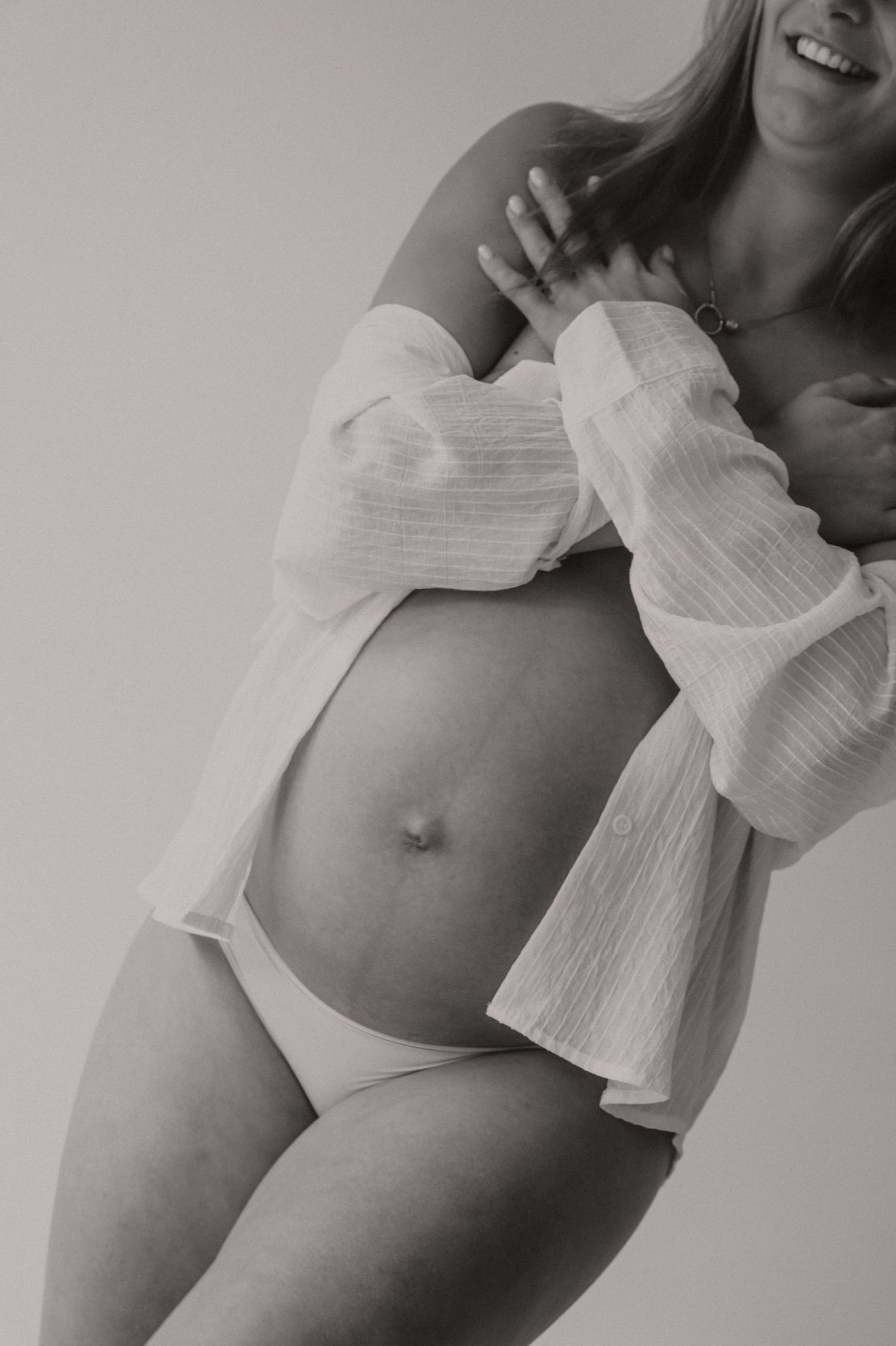 Zwangere vrouw in witte lingerie staat elegant van opzij, fine art zwangerschapsfotografie Roeselare