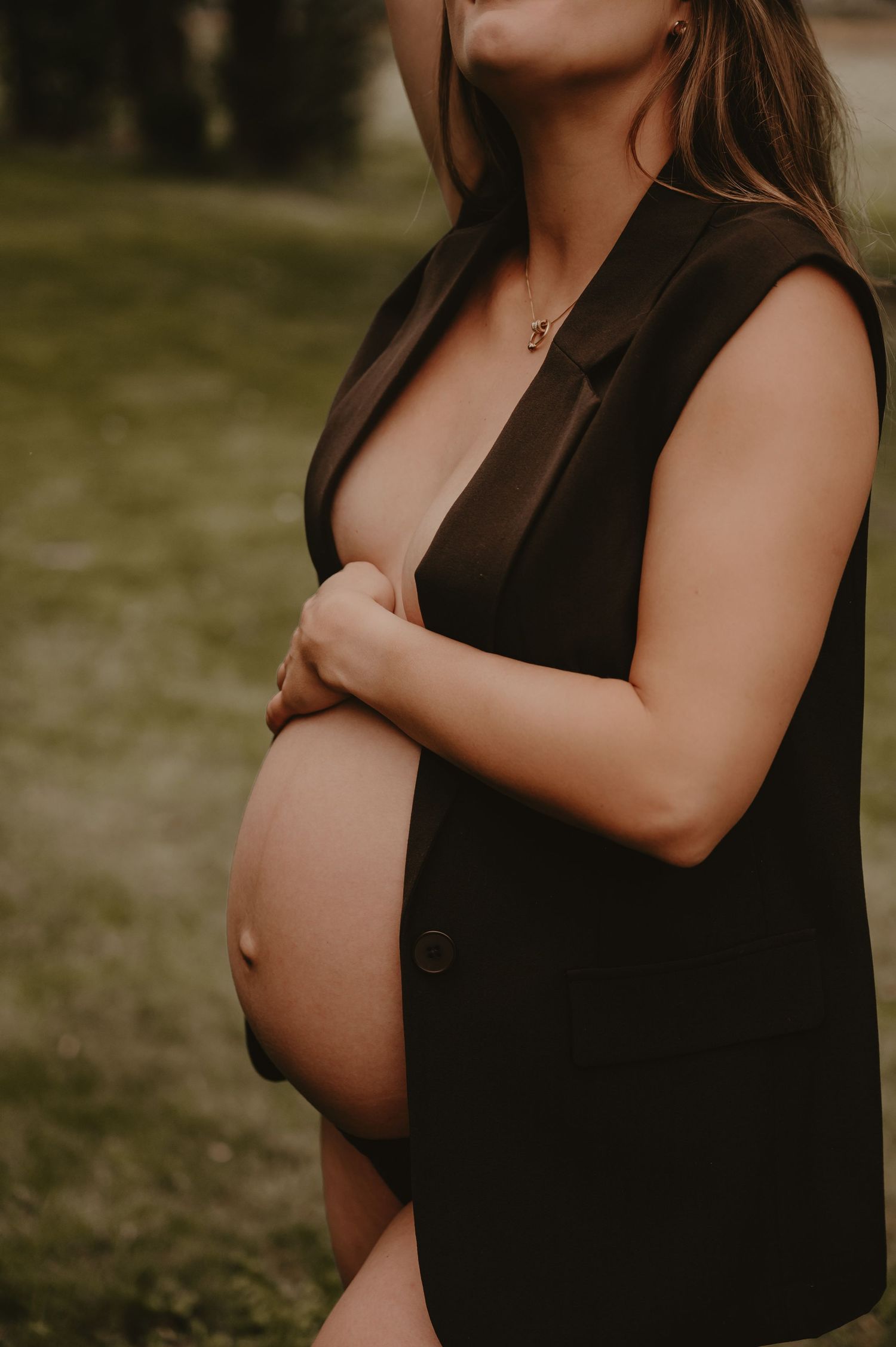 Zwangere vrouw in zwarte jurk houdt haar buik vast in een groene buitenomgeving, zwangerschapsfotograaf Roeselare