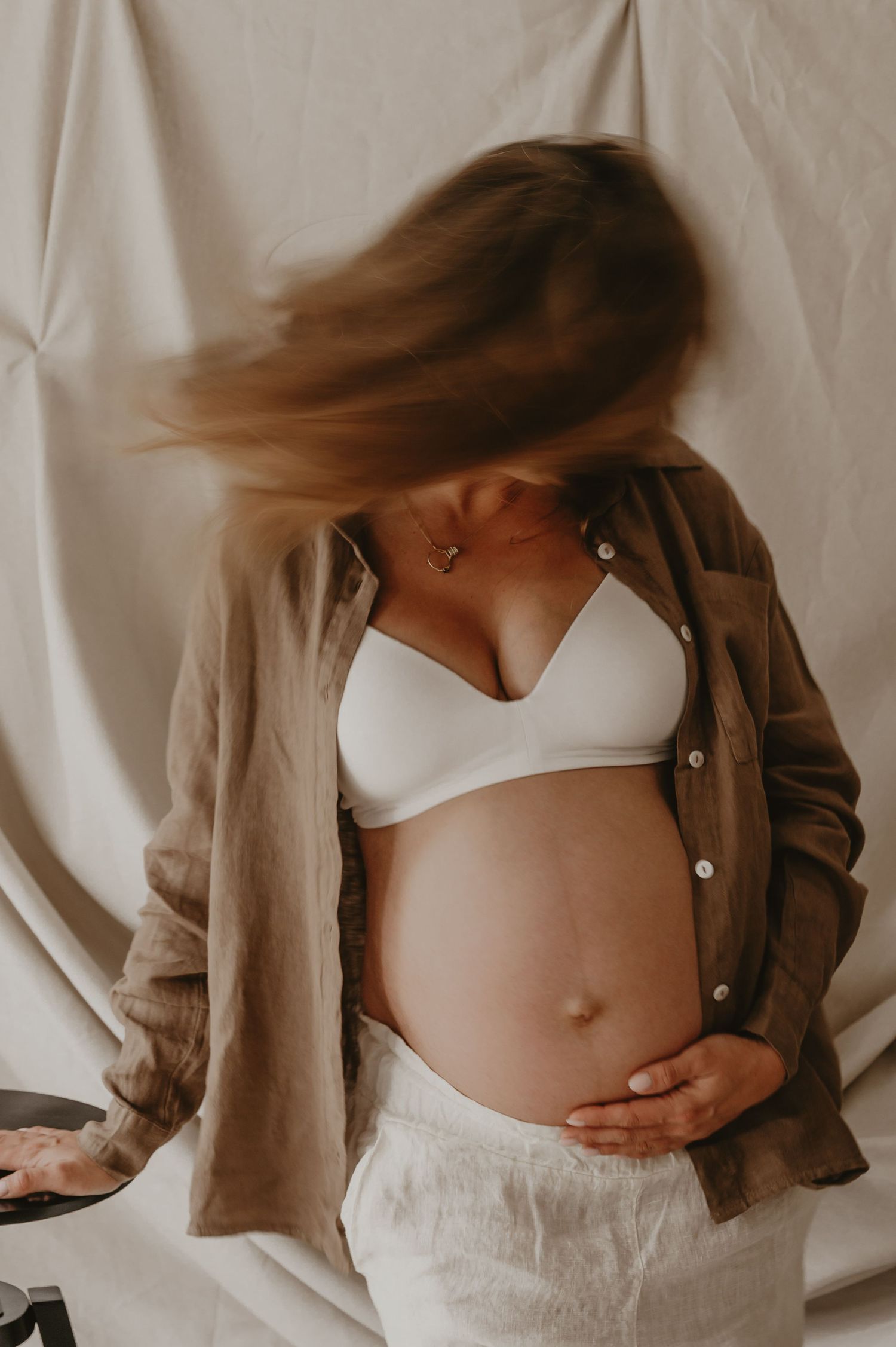 Zwangere vrouw met wapperende haren houdt buik vast in beige cardigan, lifestyle zwangerschapsfotografie Roeselare
