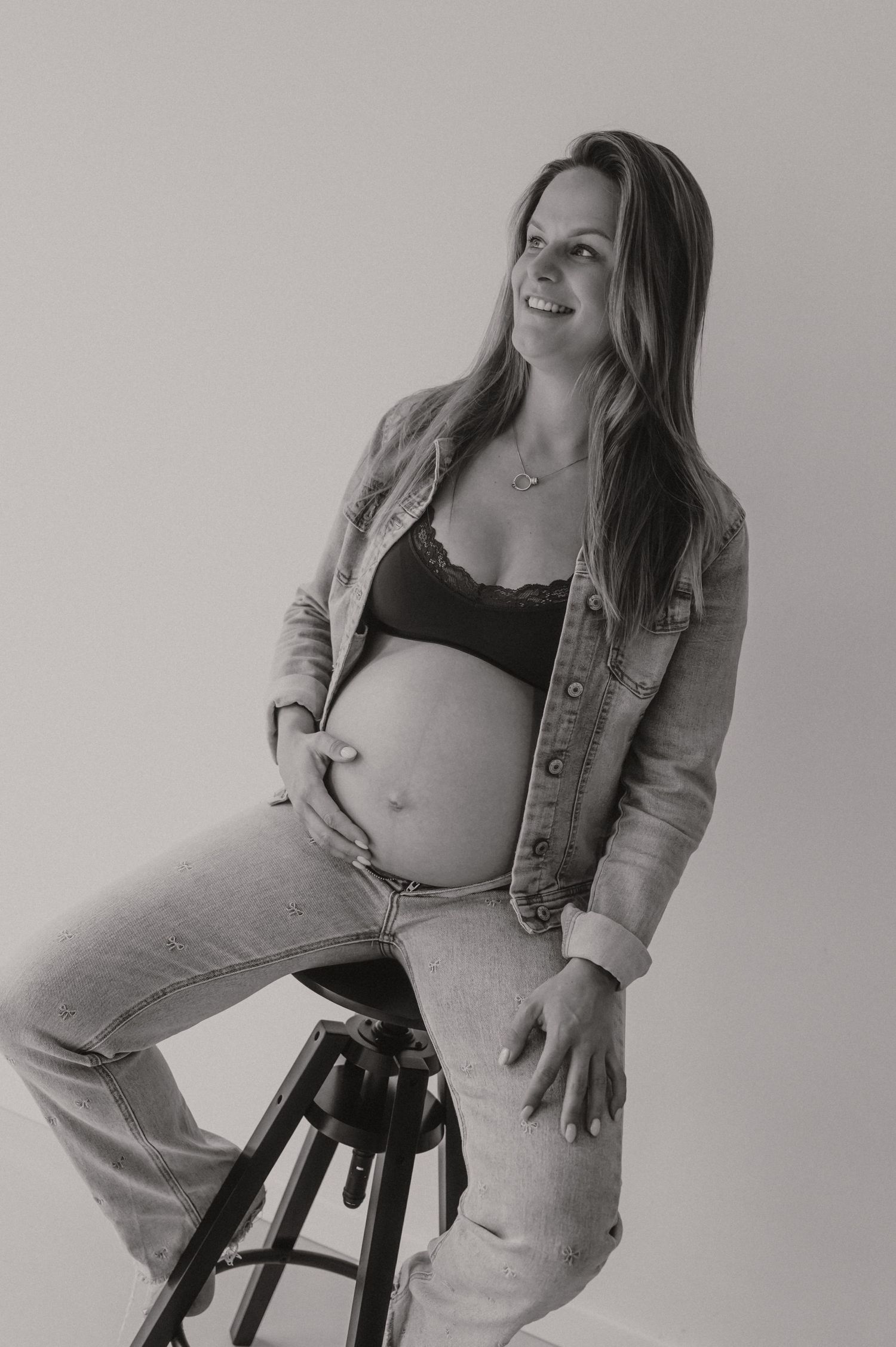 Zwangere vrouw in zwart-wit zit op kruk in studio, elegante zwangerschapsfotografie fotograaf Roeselare