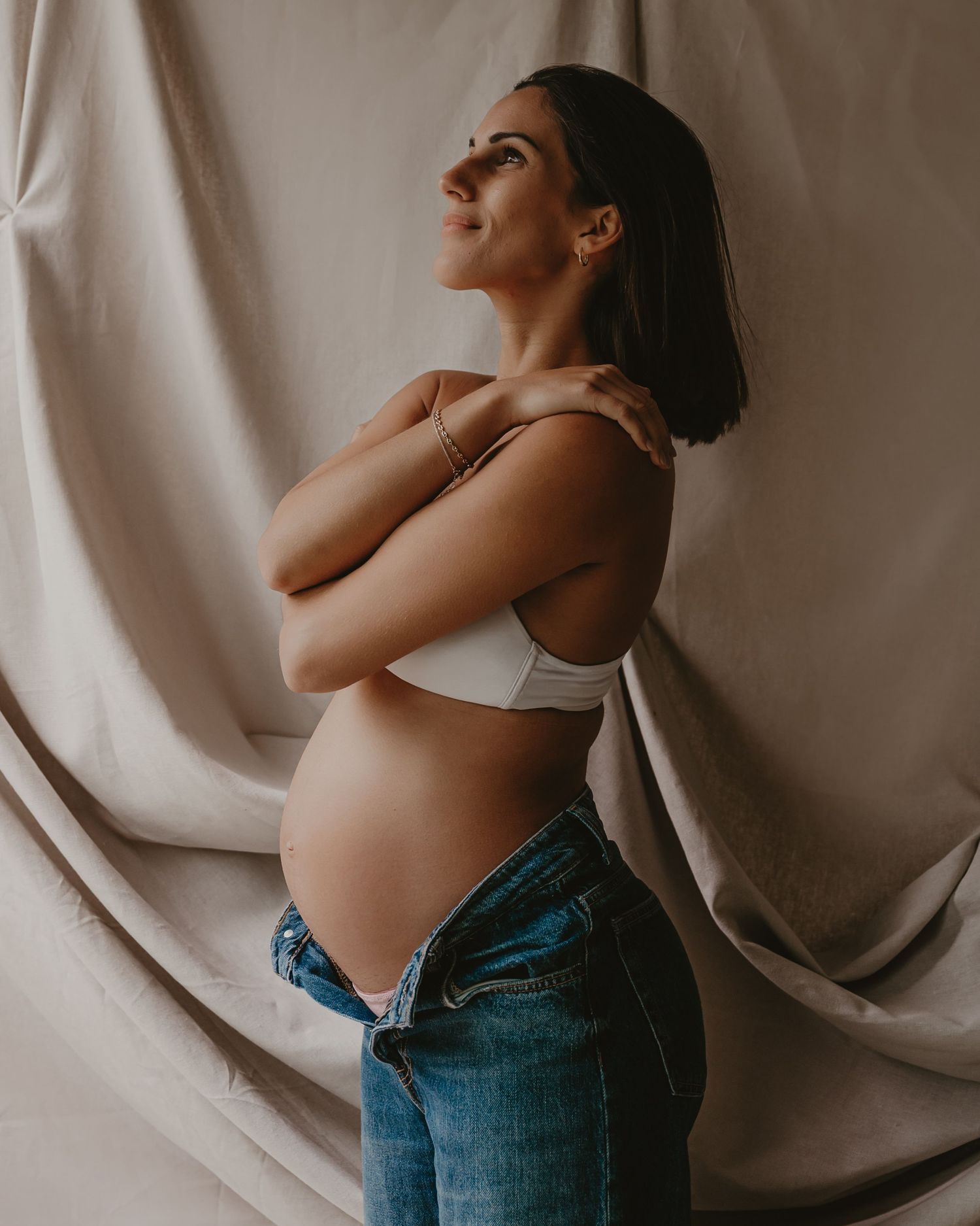 Zwangere vrouw kijkt omhoog in studio met linnen gordijn, fine art zwangerschapsfotografie Roeselare