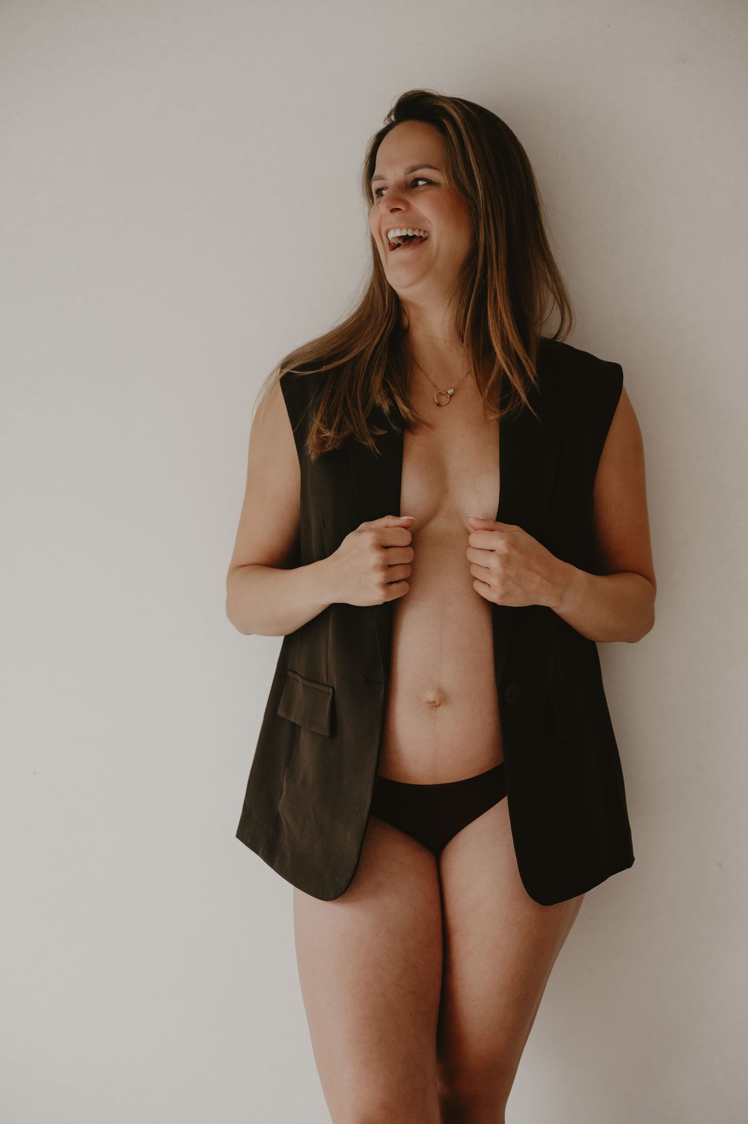 Zwangere vrouw in donker linnen vest poseert in studio met witte achtergrond, zwangerschapsfotograaf Roeselare