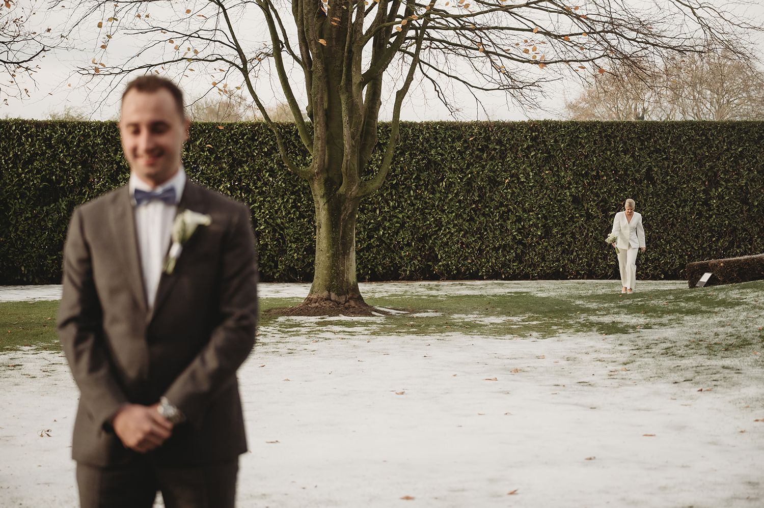Bruidegom wacht in besneeuwde tuin terwijl de bruid nadert — huwelijksfotograaf West-Vlaanderen