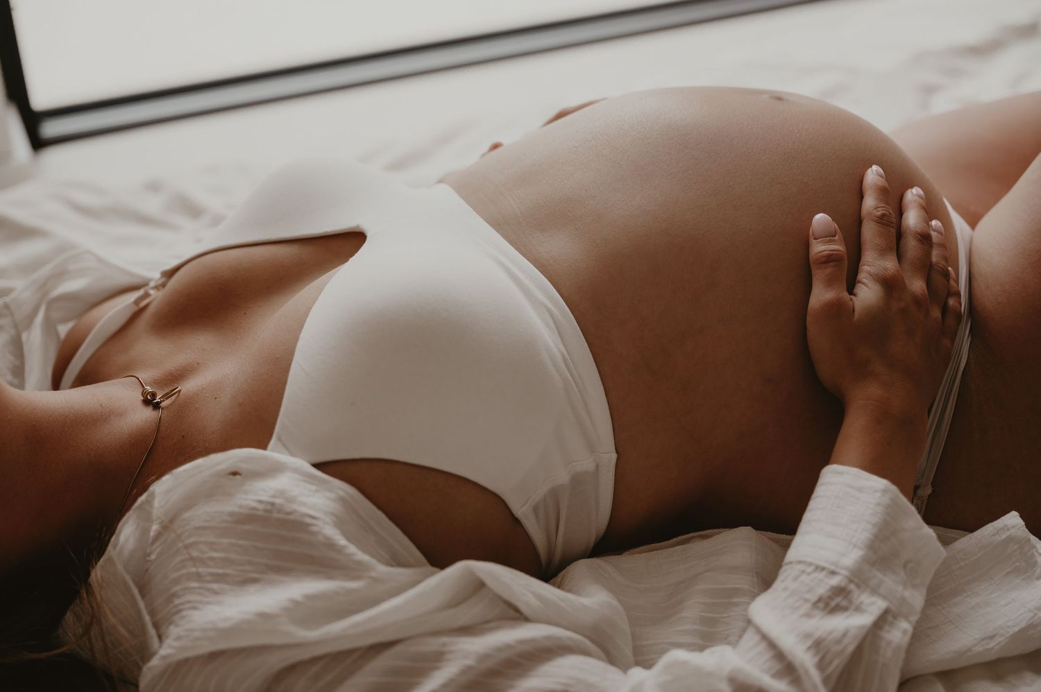 Zwangere vrouw ligt met warme belichting en hand op buik, sensuele zwangerschapsfotografie Roeselare