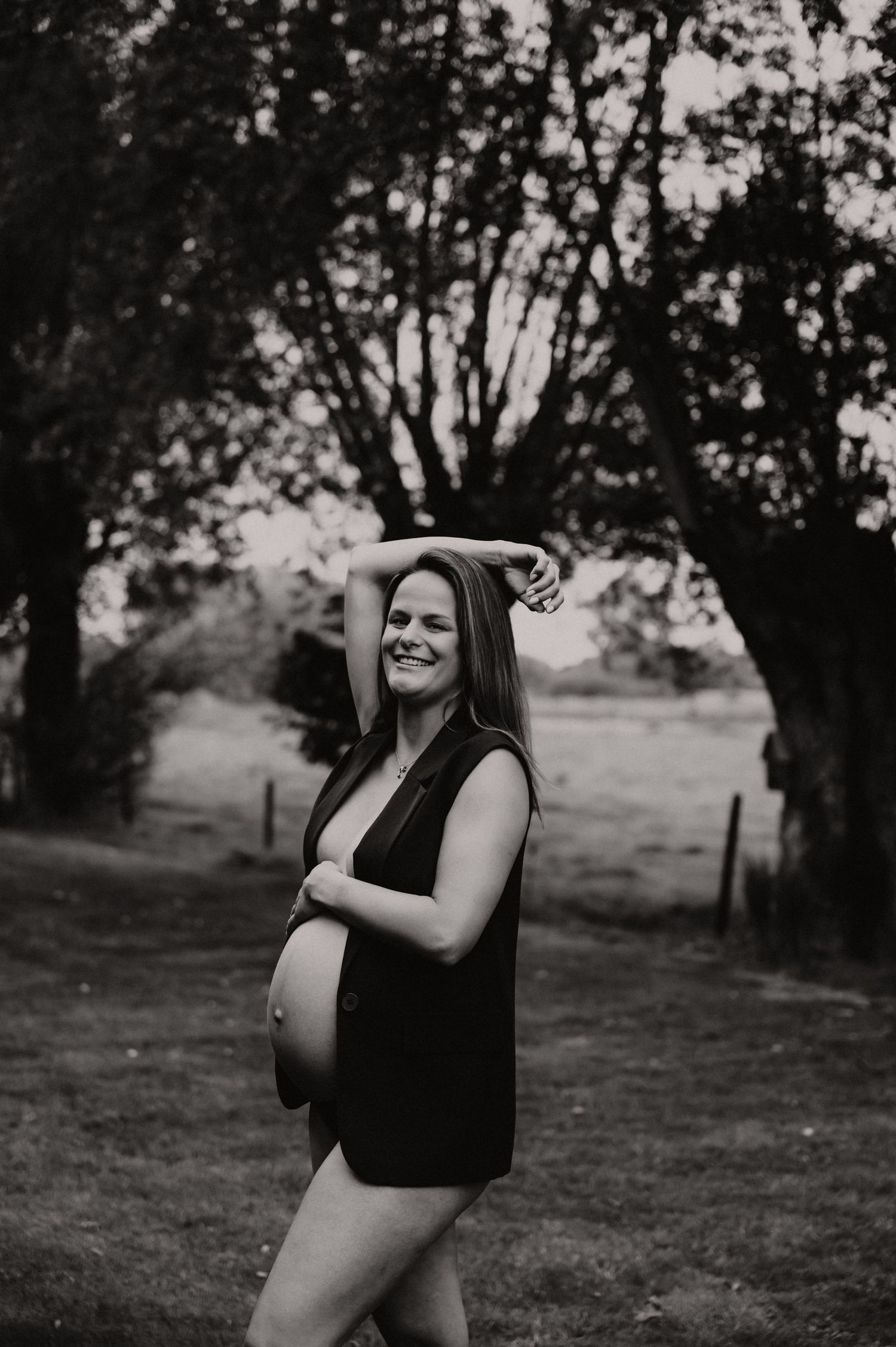 Zwangere vrouw lacht vrolijk buiten in zwart-wit met donker vest, lifestyle zwangerschapsfotografie West-Vlaanderen