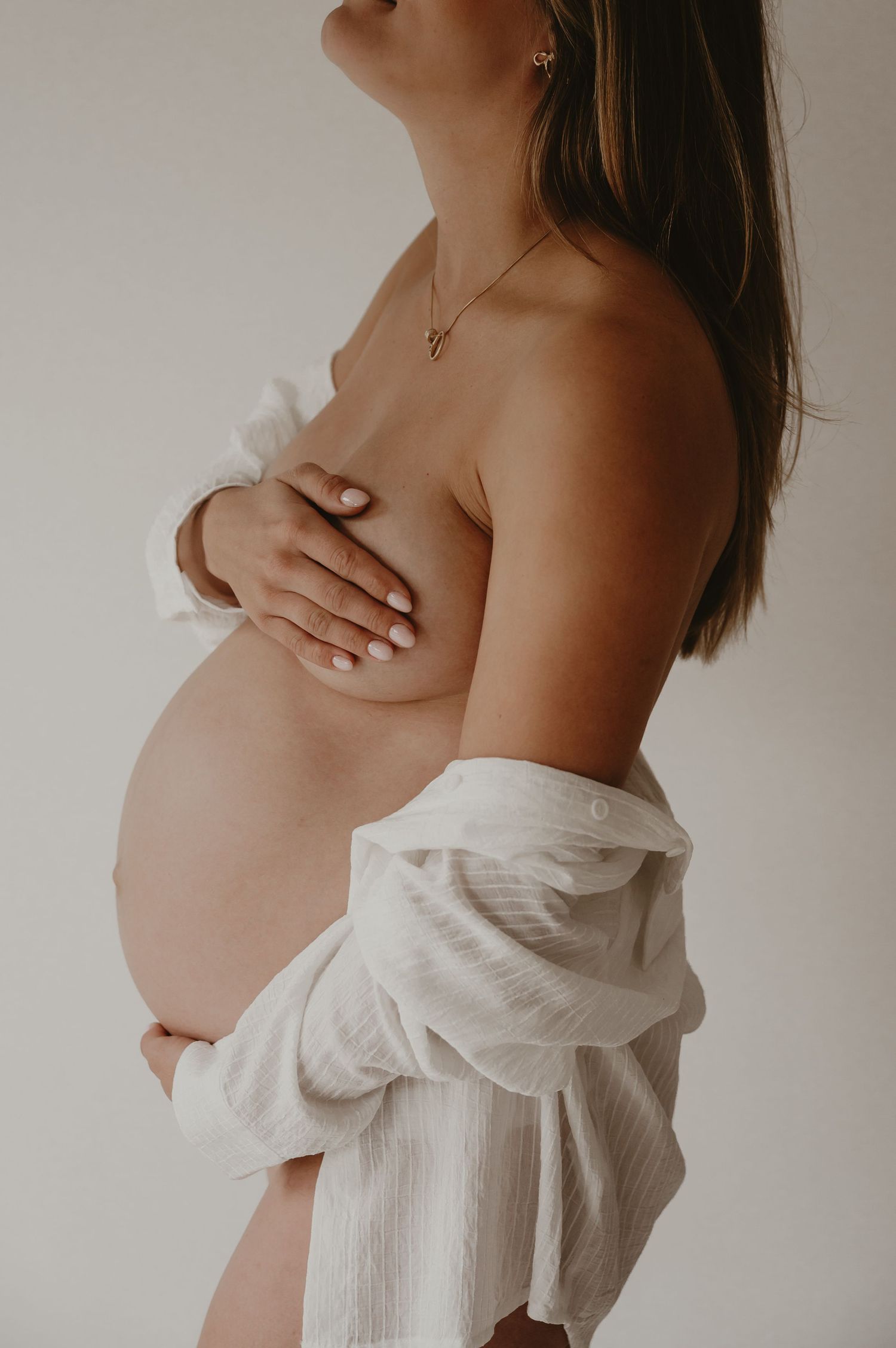 Zwangere vrouw in stromende witte jurk houdt buik vast, elegante zwangerschapsfotografie Roeselare