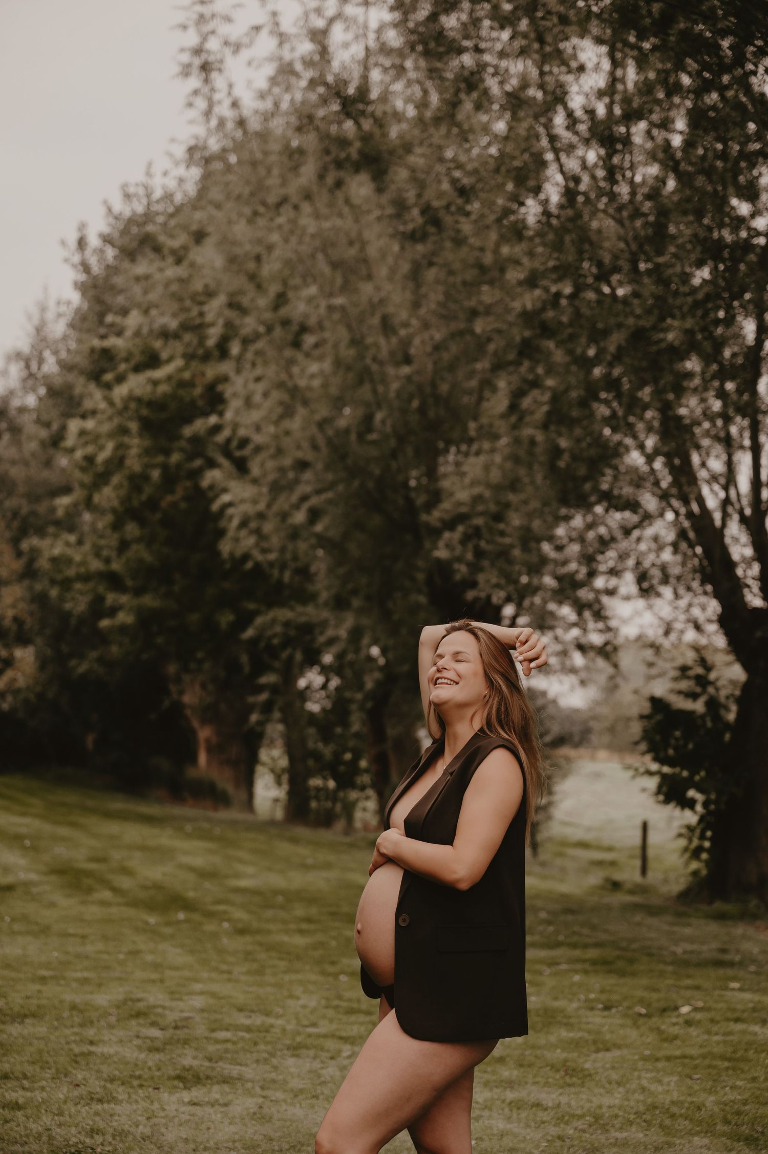 Zwangere vrouw lacht en speelt met haar haren in een groene tuin, outdoor zwangerschapsfotografie Roeselare