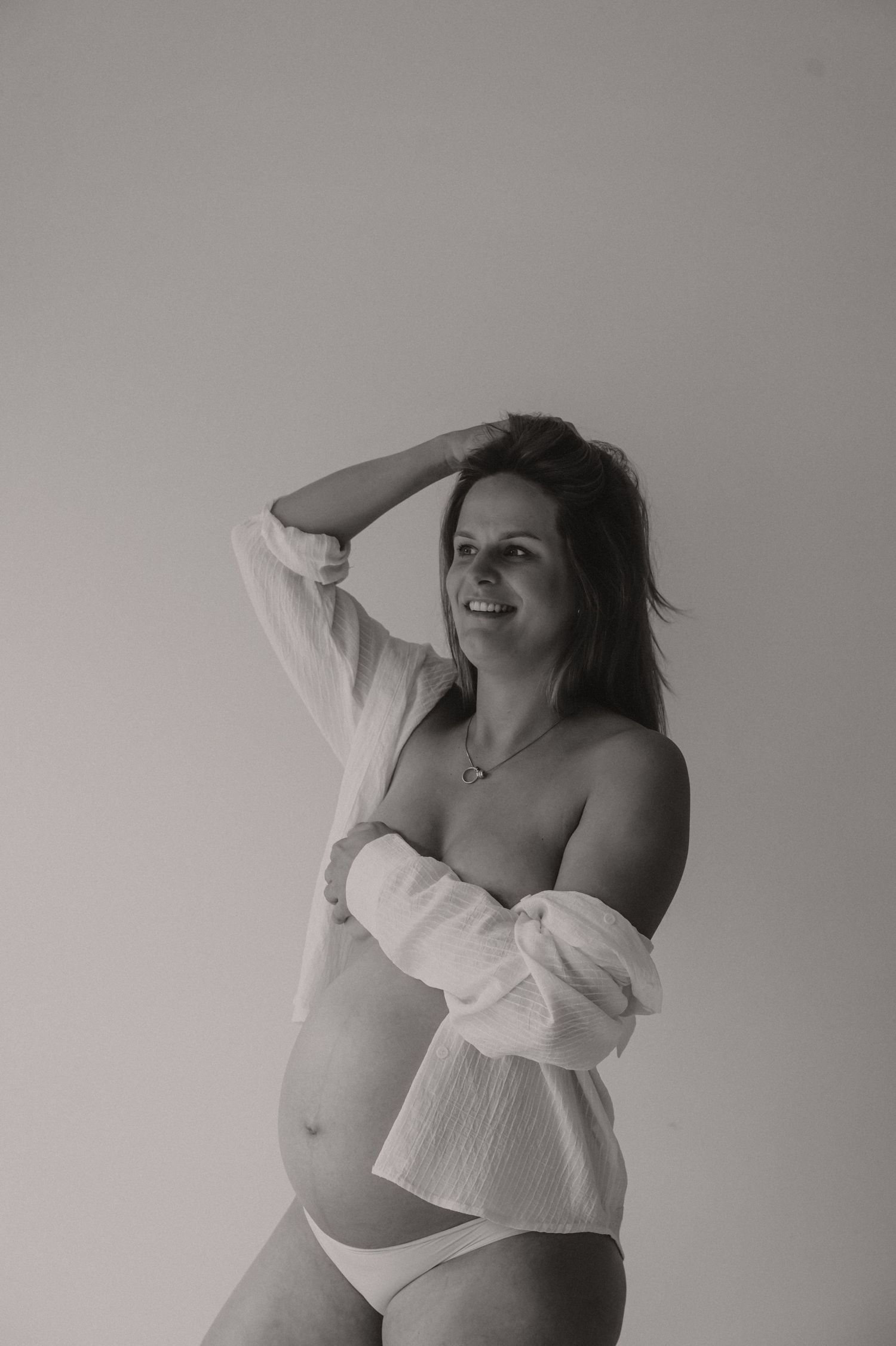 Zwangere vrouw in witte lingerie poseert sierlijk met armen, fine art zwangerschapsfotografie Roeselare