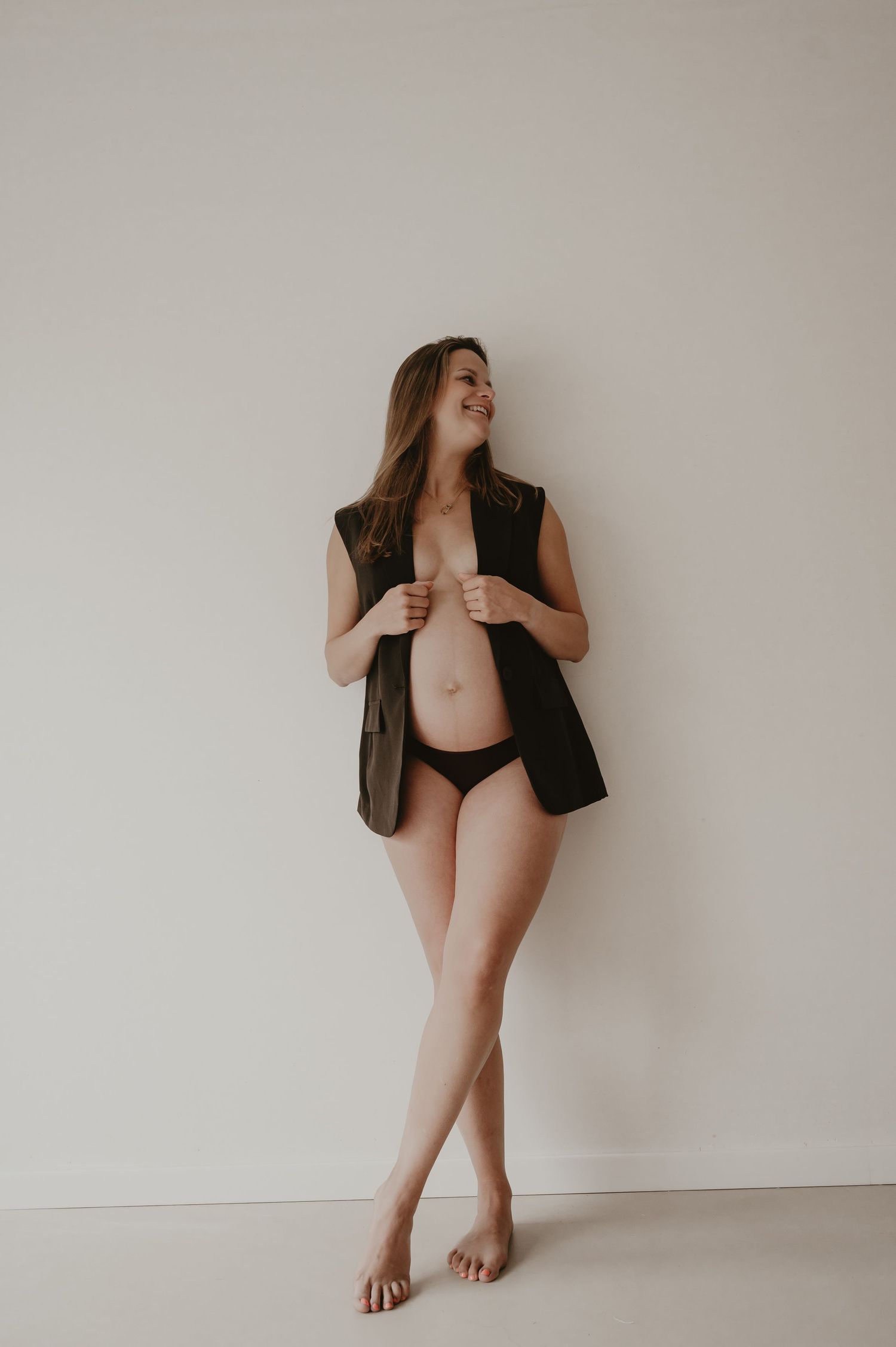 Zwangere vrouw in lang zwart vest poseert elegant in studio, zwangerschapsfotograaf Roeselare