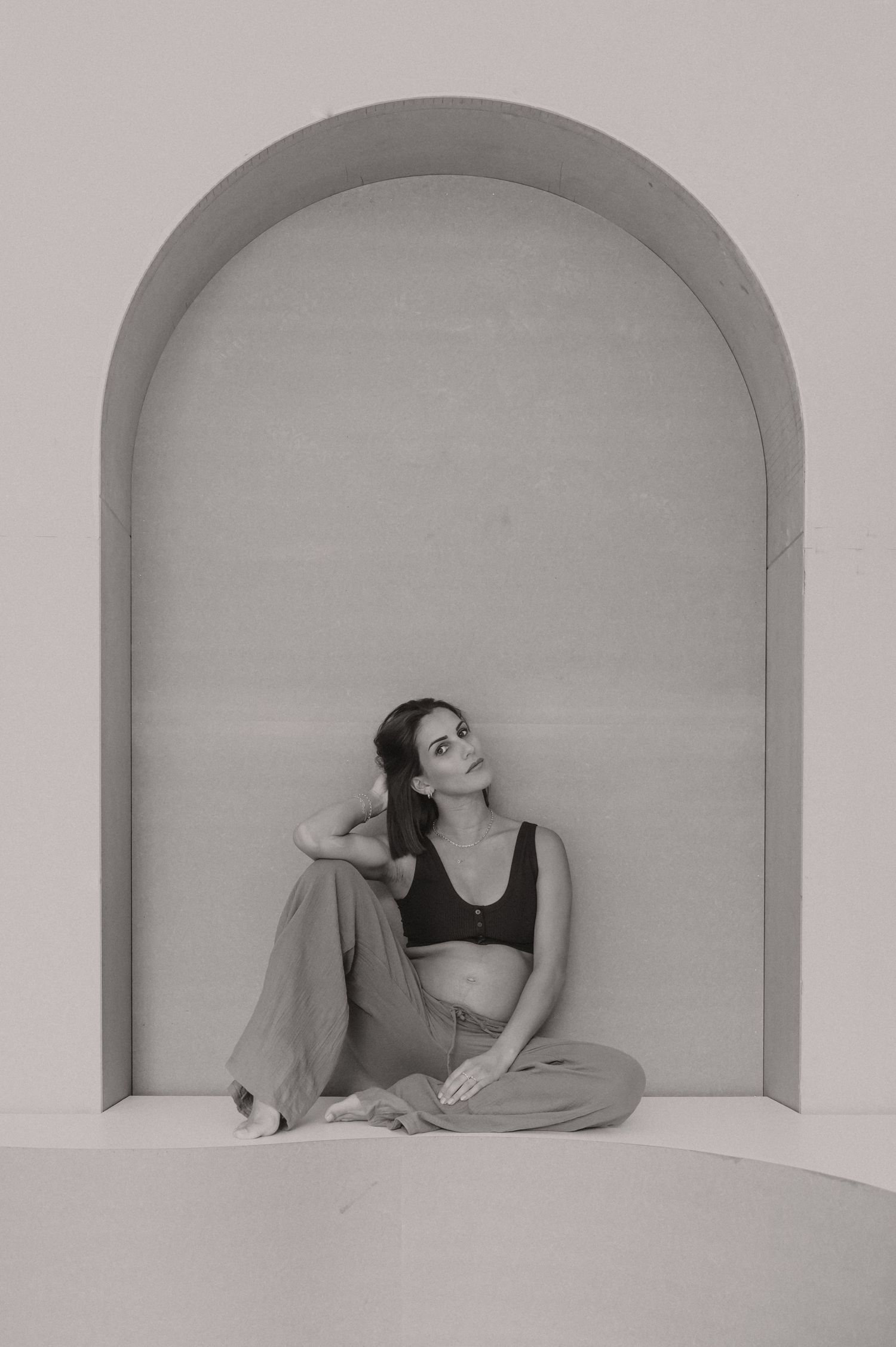 Zwangere vrouw zit kalm in boogvormige nis, zwart-wit zwangerschapsfotografie minimalistische studio Roeselare