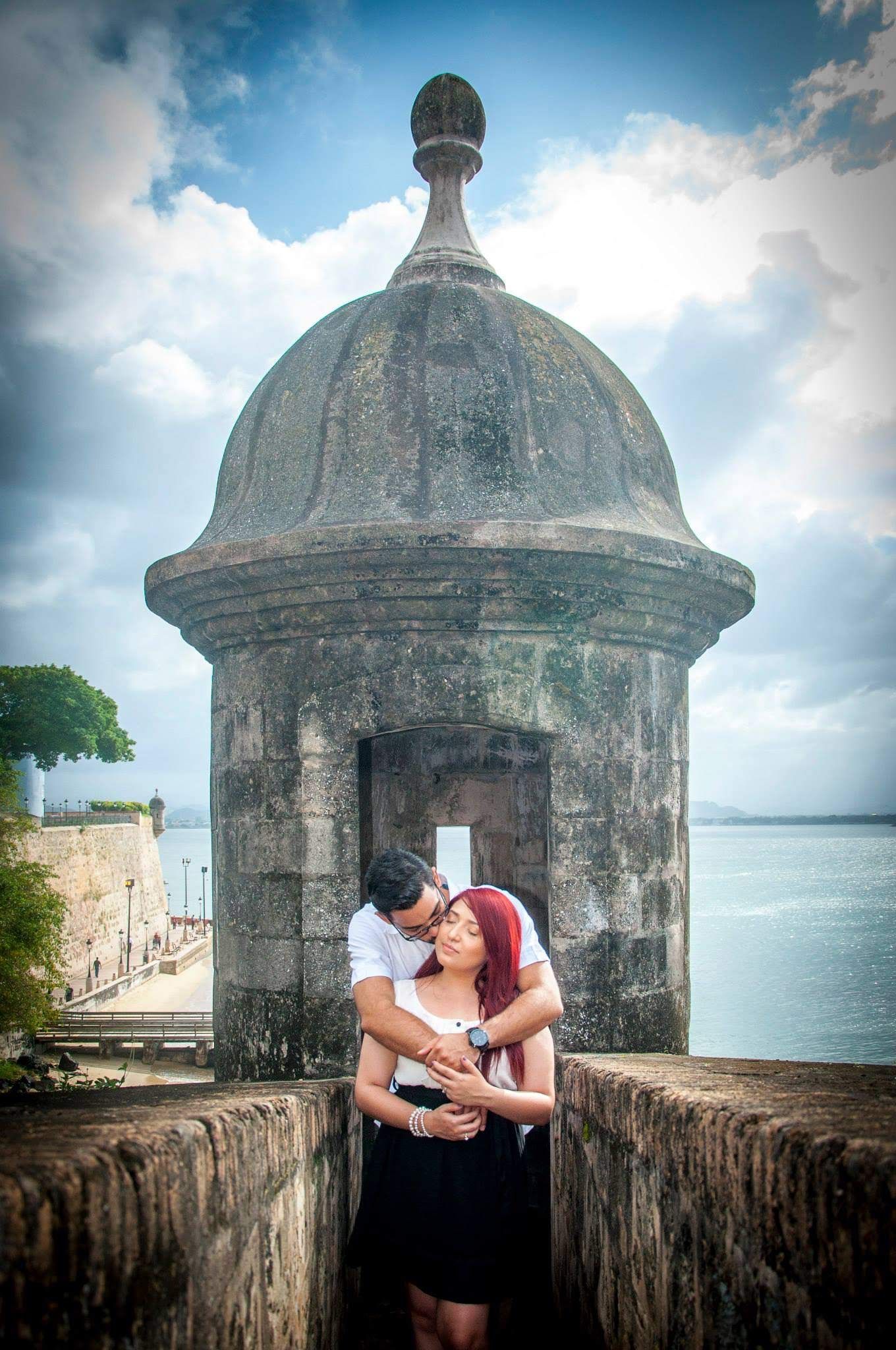 A couple embraces in front of a historic stone guard tower with ocean views in the background.