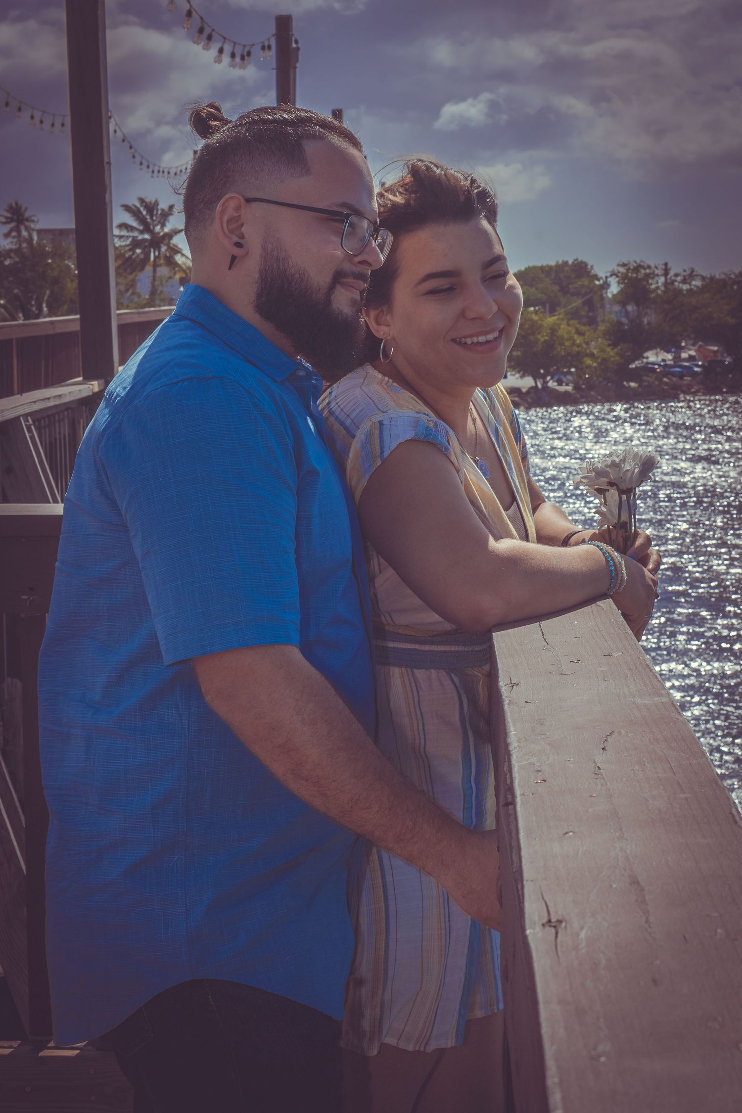 A romantic couple shares an intimate moment while looking out over sparkling water from a waterfront deck on a sunny day.