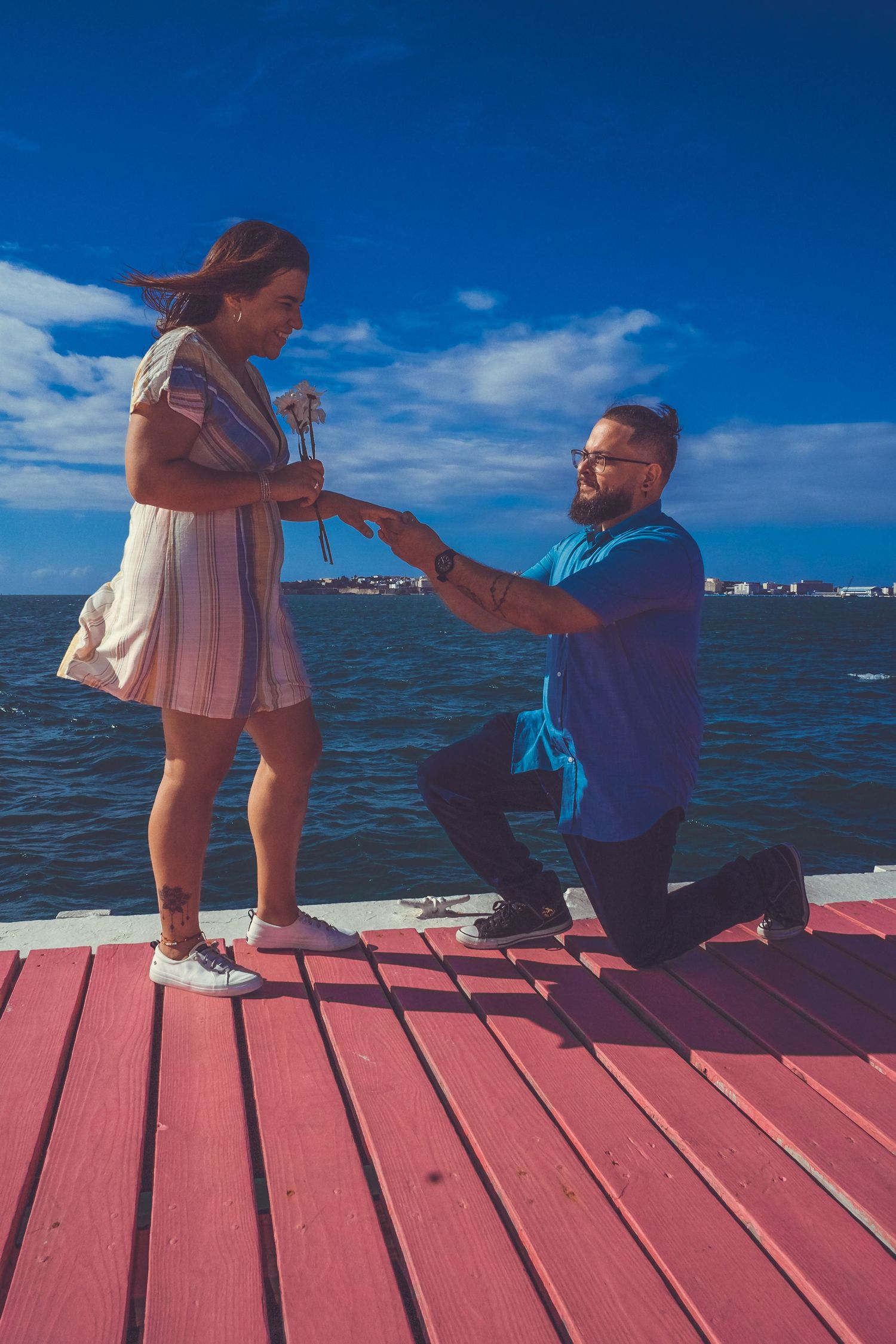 A romantic proposal scene unfolds on a red wooden pier with ocean and blue skies in the background.