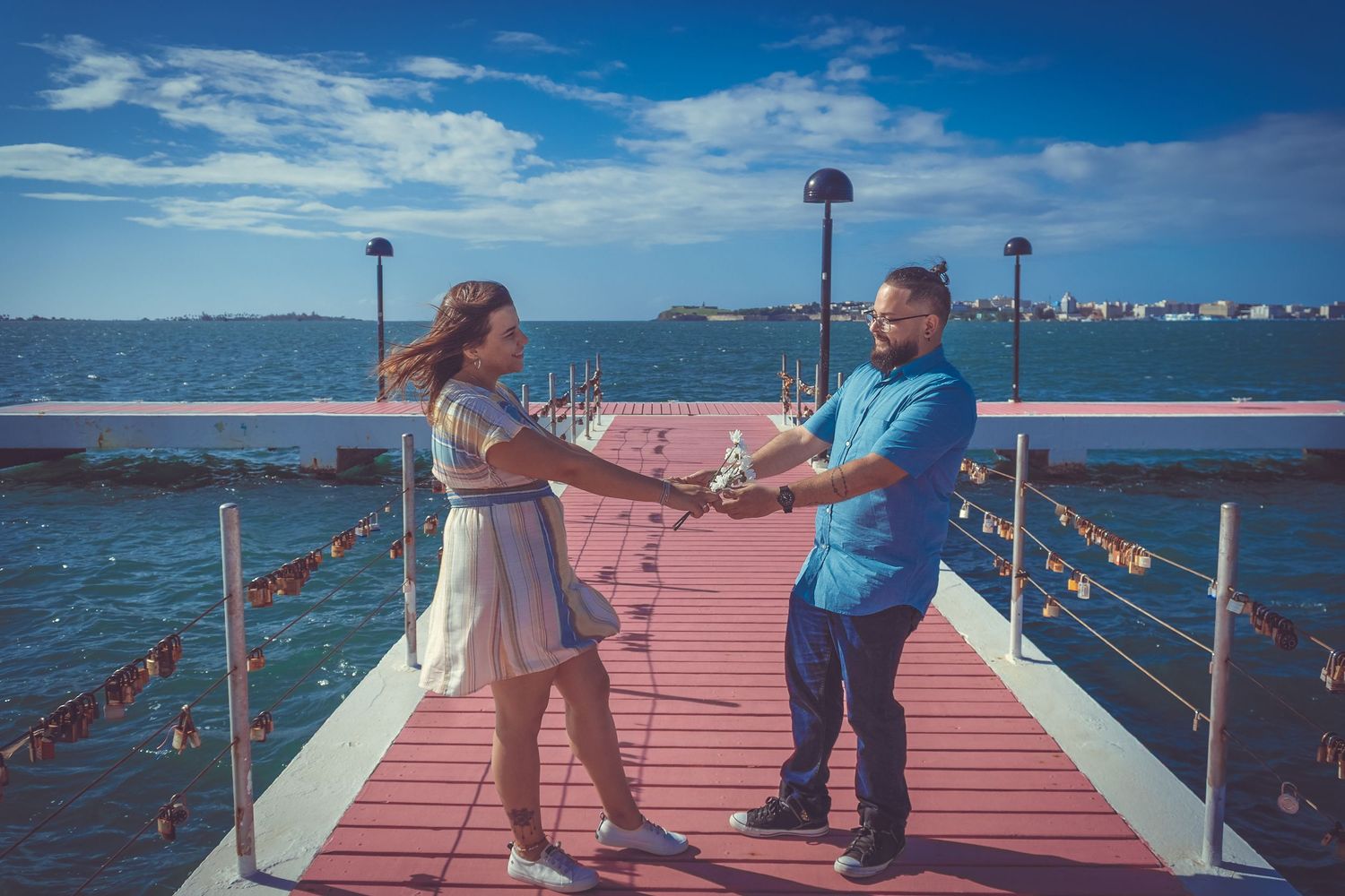 A couple holds hands while standing on a red pier walkway with water and city views in the background.
