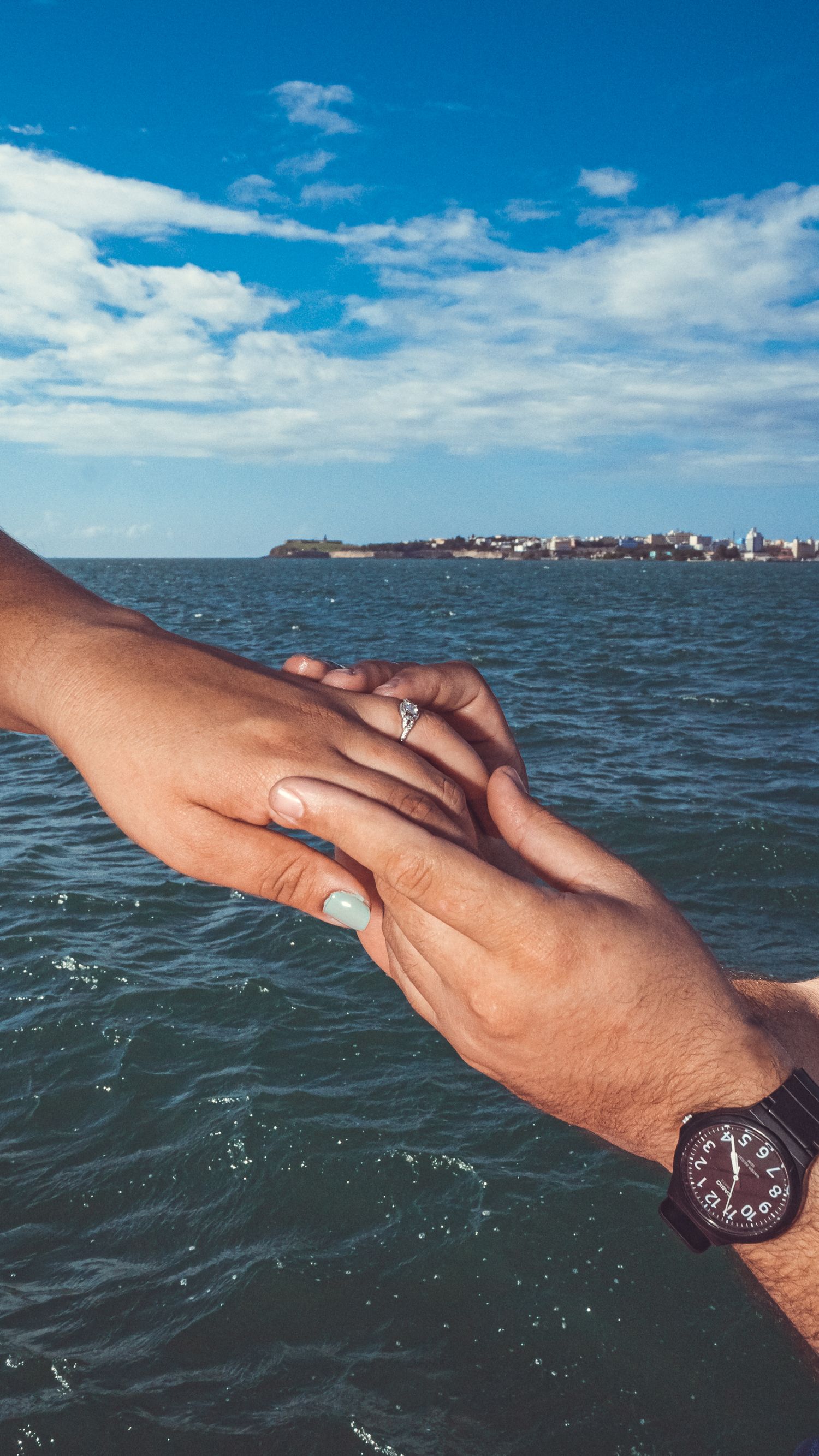 Two hands join together over ocean waters with a distant cityscape visible on the horizon.