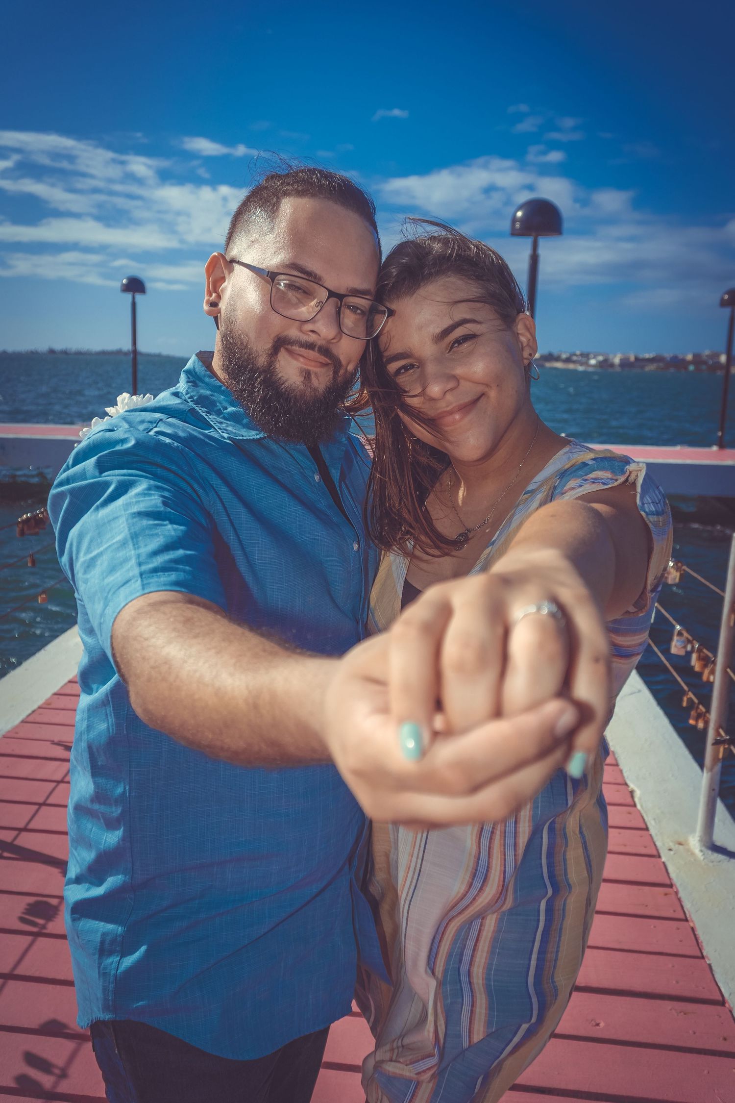 A couple shows off an engagement ring while posing on a waterfront pier under blue skies.