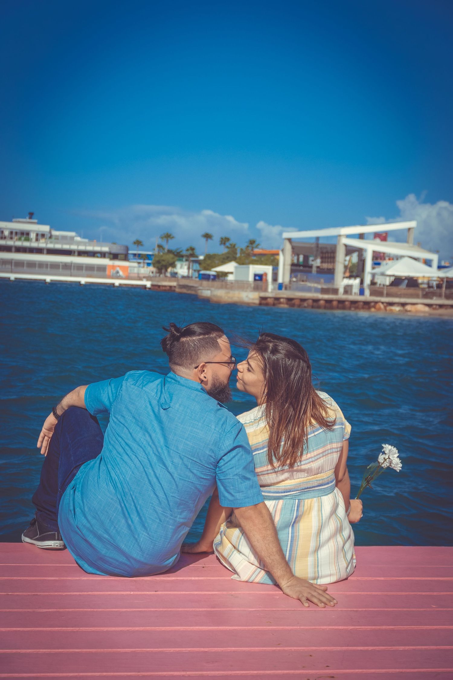 A couple sits close together on a red pier deck overlooking the ocean and coastal buildings.