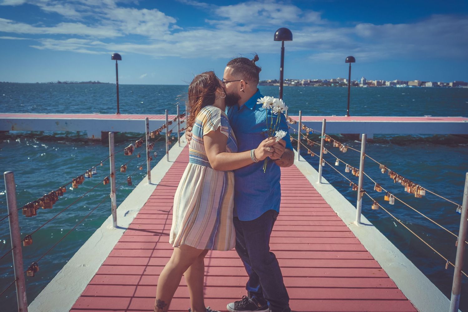 A couple embraces on a red waterfront pier with lamp posts and ocean waters surrounding them.